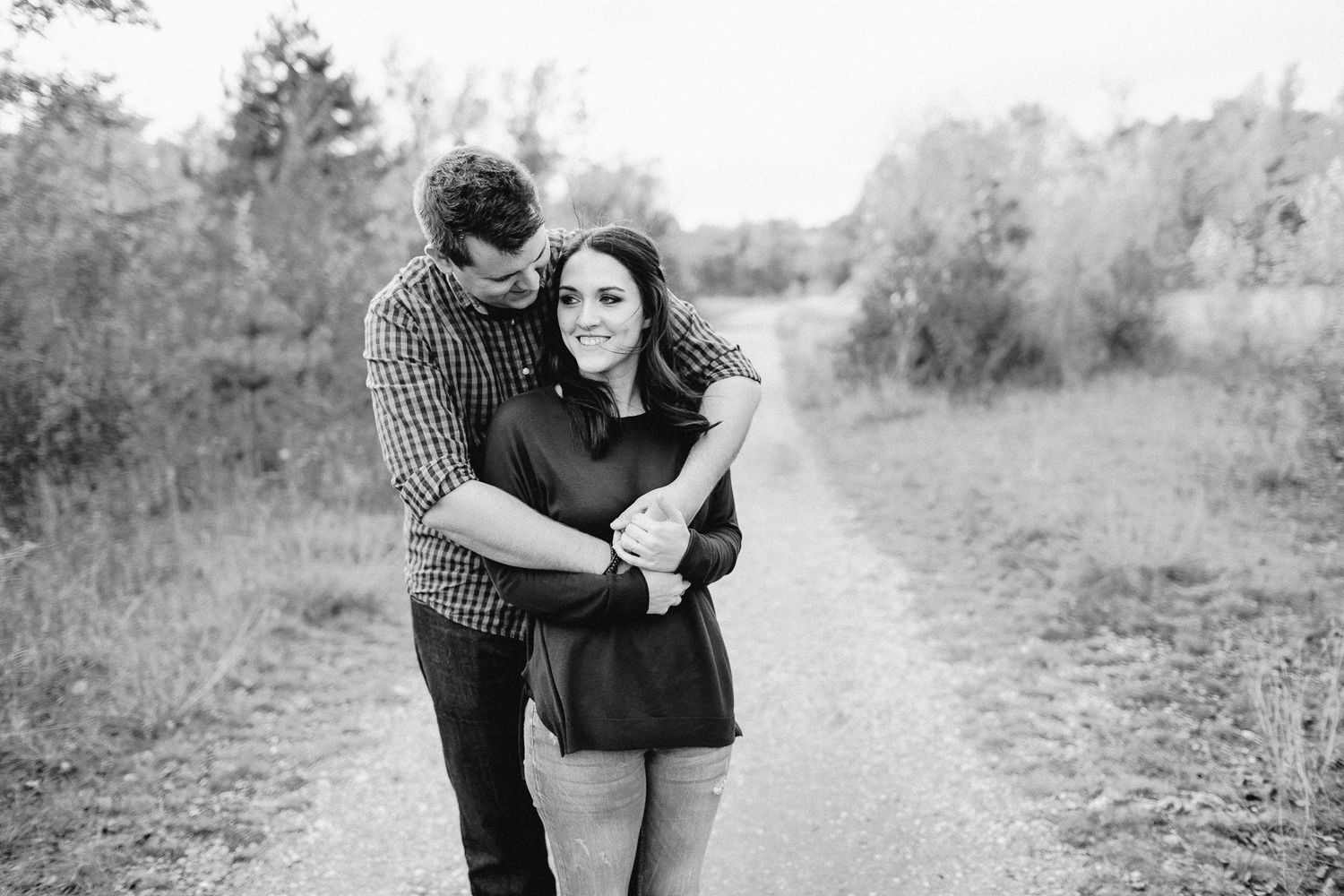 Black and white photo of a couple embracing on a scenic outdoor path surrounded by tall grass.