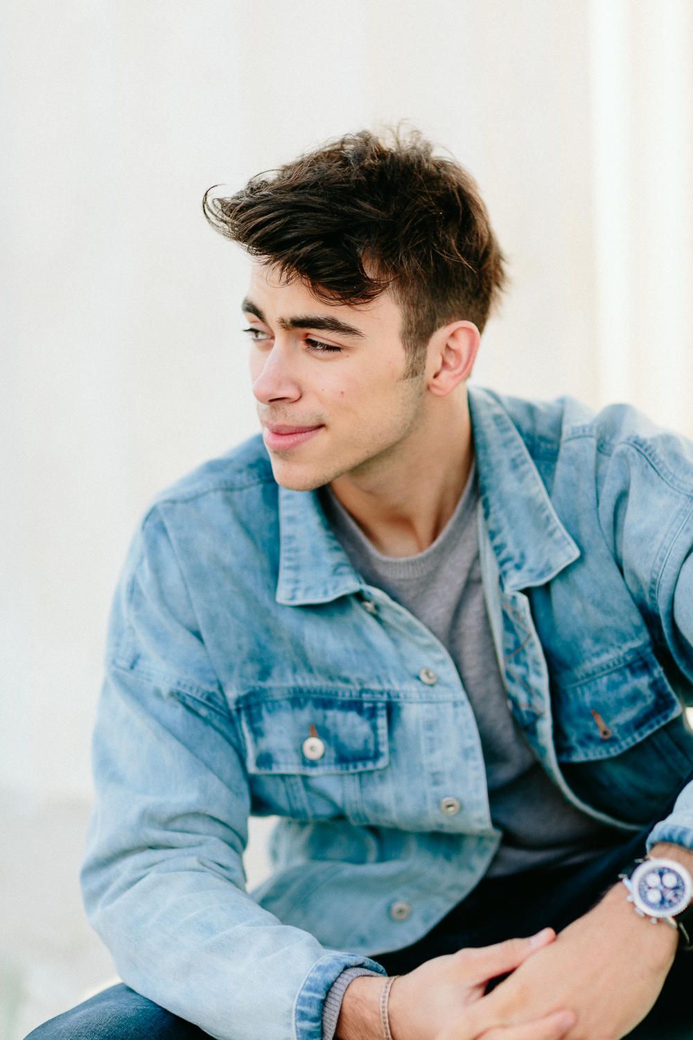 Young man in denim jacket looking sideways wearing a watch in casual outdoor portrait.