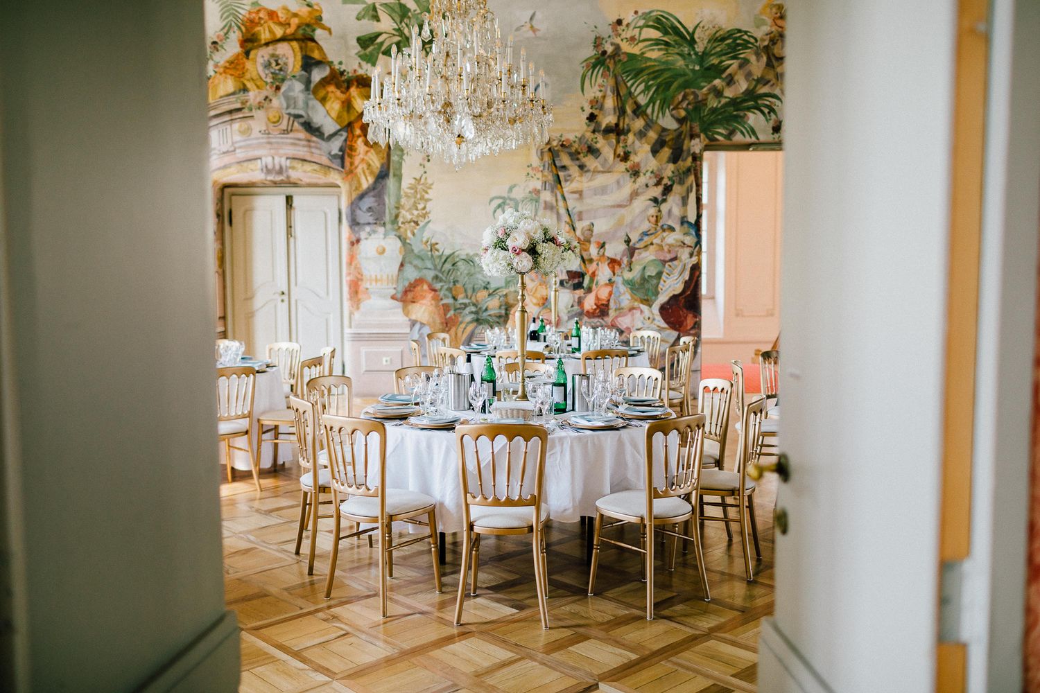 Elegant wedding reception room with round tables, gold chairs, chandelier, and ornate floral mural wallpaper.