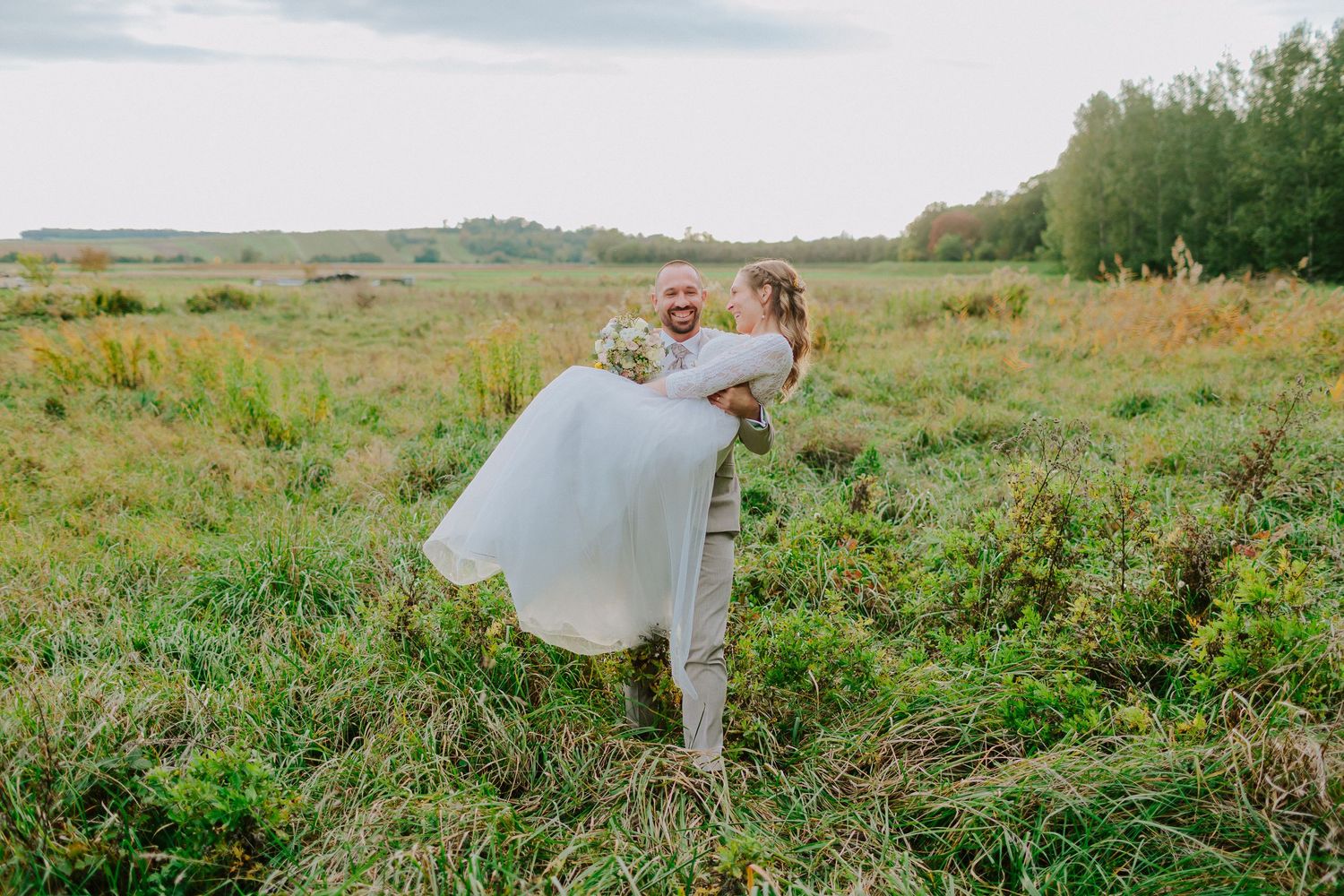 Groom lifting bride in white dress while laughing together in a lush green meadow during outdoor wedding.