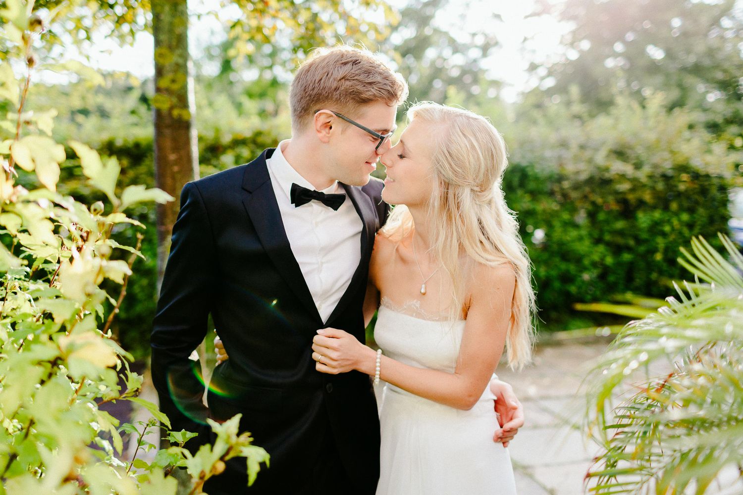 Blonde bride and groom in black tuxedo sharing intimate almost-kiss surrounded by lush green garden foliage.