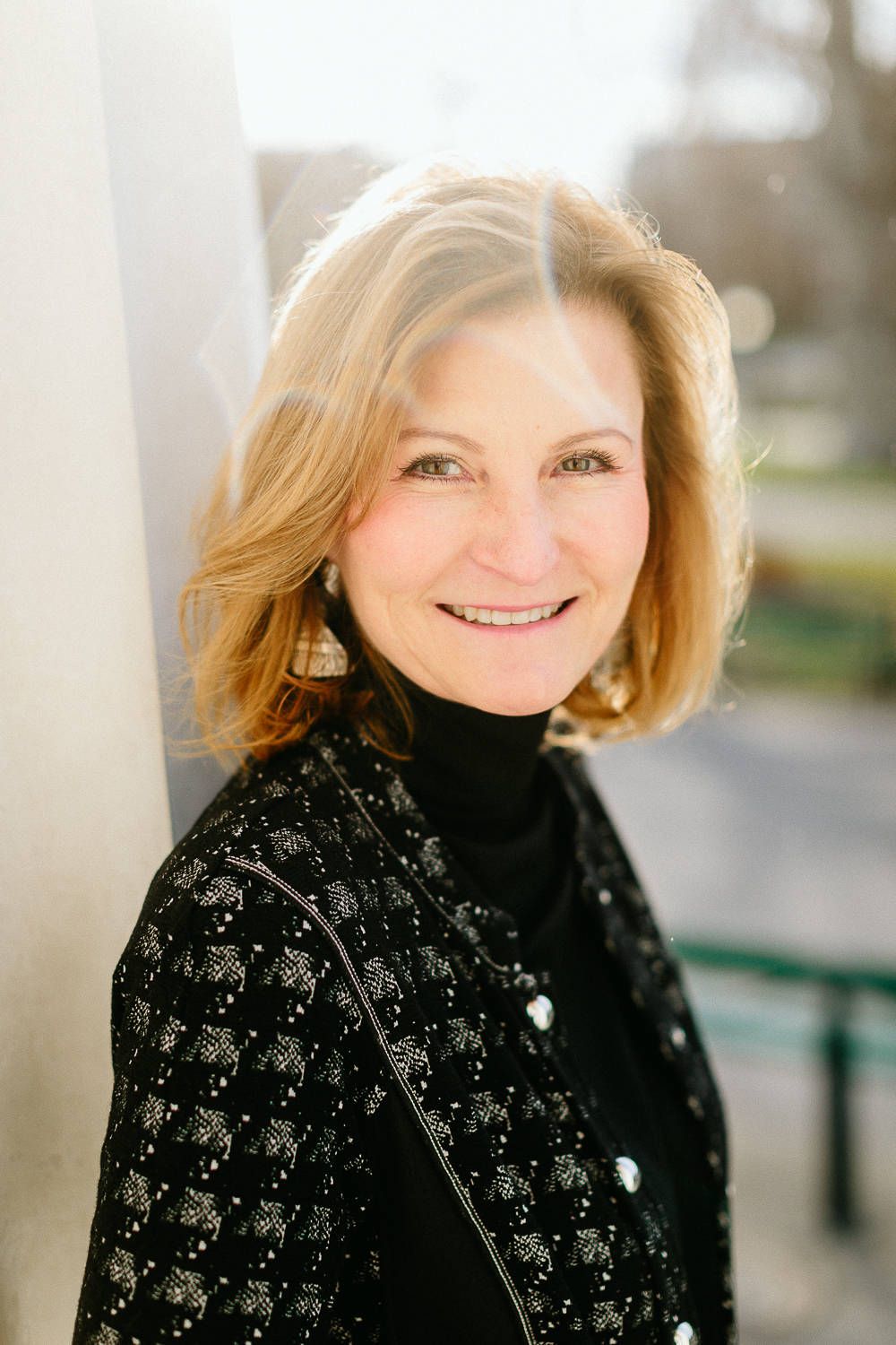 Smiling mature woman with auburn hair in black sequined jacket leaning against white column.