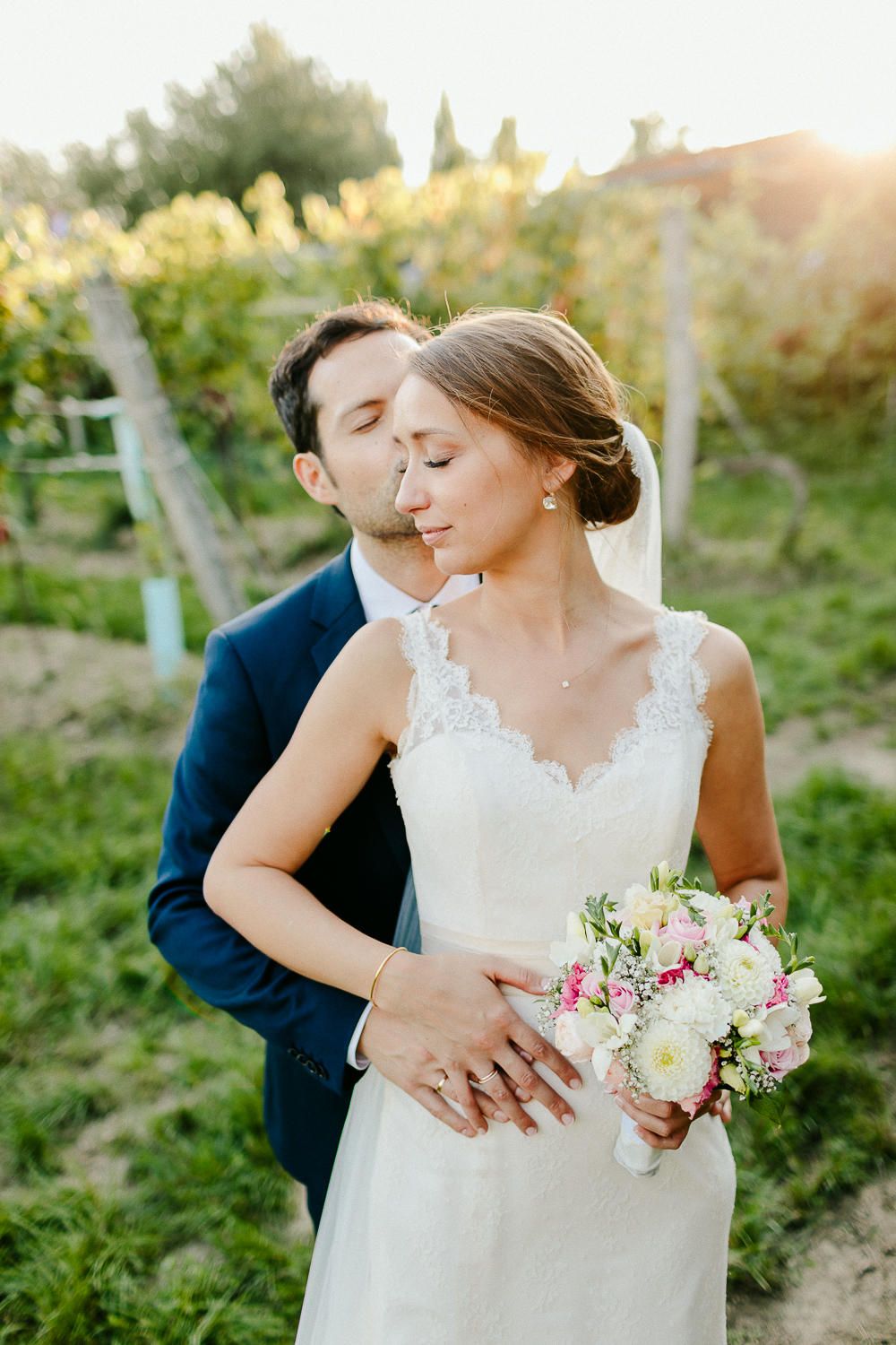 Bride and groom share a tender moment in a sunlit vineyard, bride holding a pink and white bouquet.