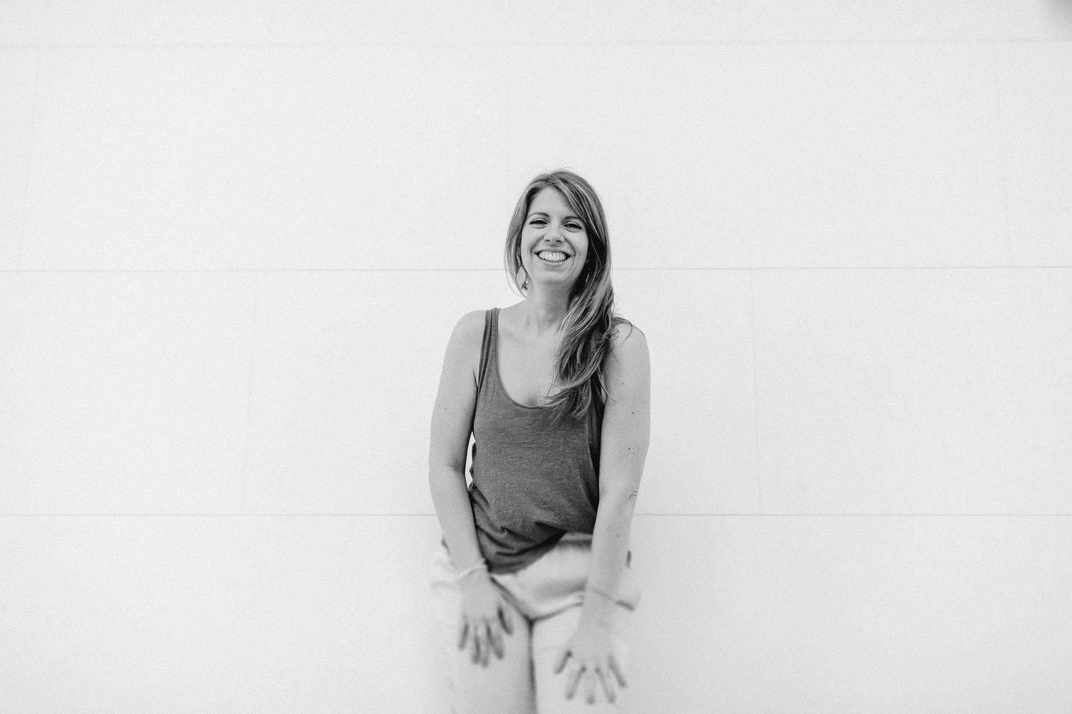 Laughing woman in gray tank top sitting casually against white wall in black and white photo.