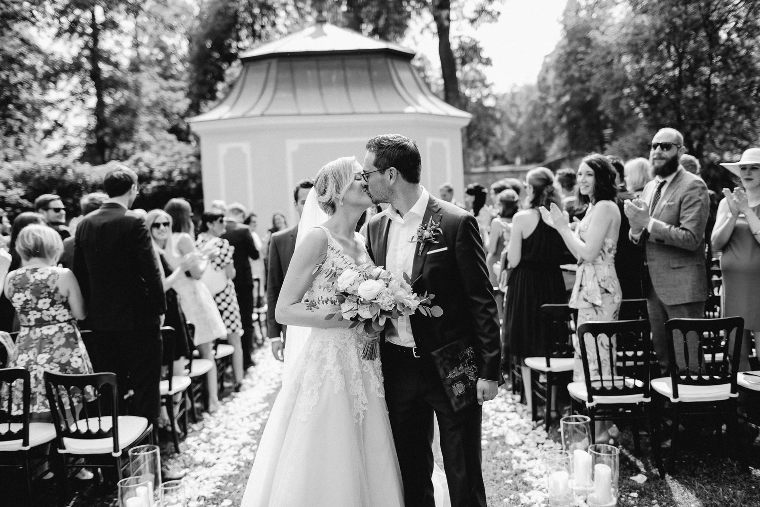 Bride and groom share first kiss at outdoor wedding ceremony with guests seated in rows.