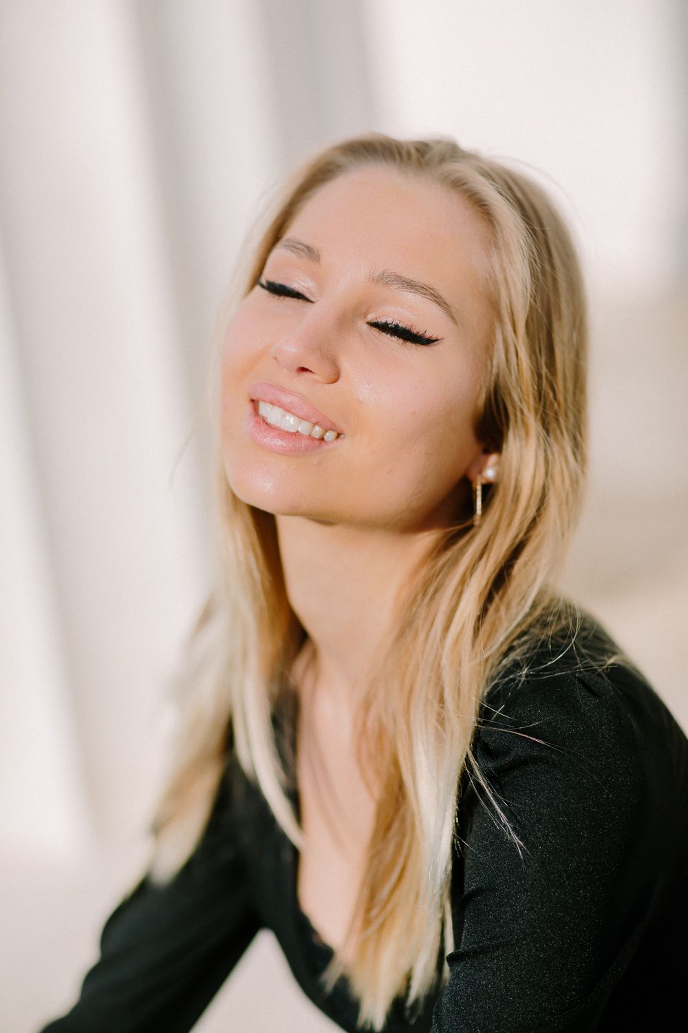 Smiling blonde woman with closed eyes, wearing black top with pearl earring in soft light.