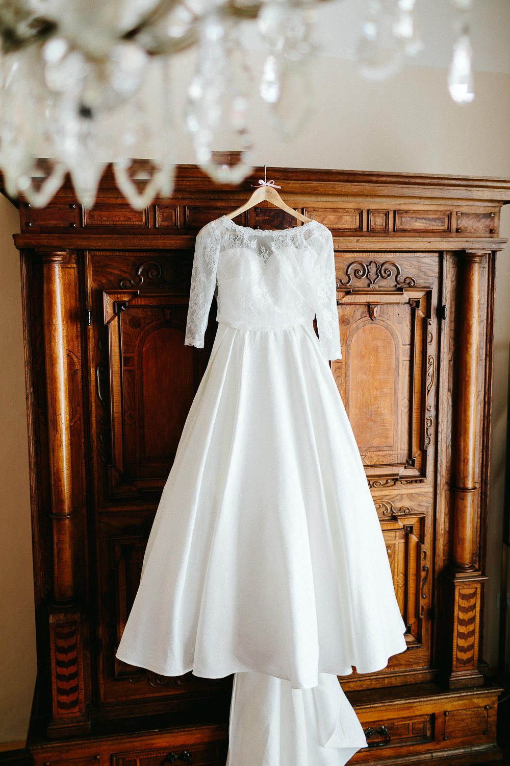 Elegant white lace wedding gown with three-quarter sleeves hanging on antique wooden wardrobe under chandelier.