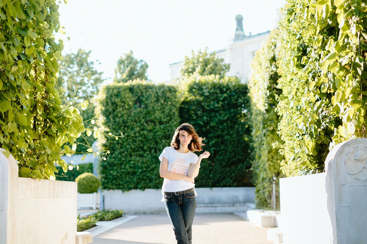 Young brunette woman walking joyfully through lush green garden pathway on sunny day.