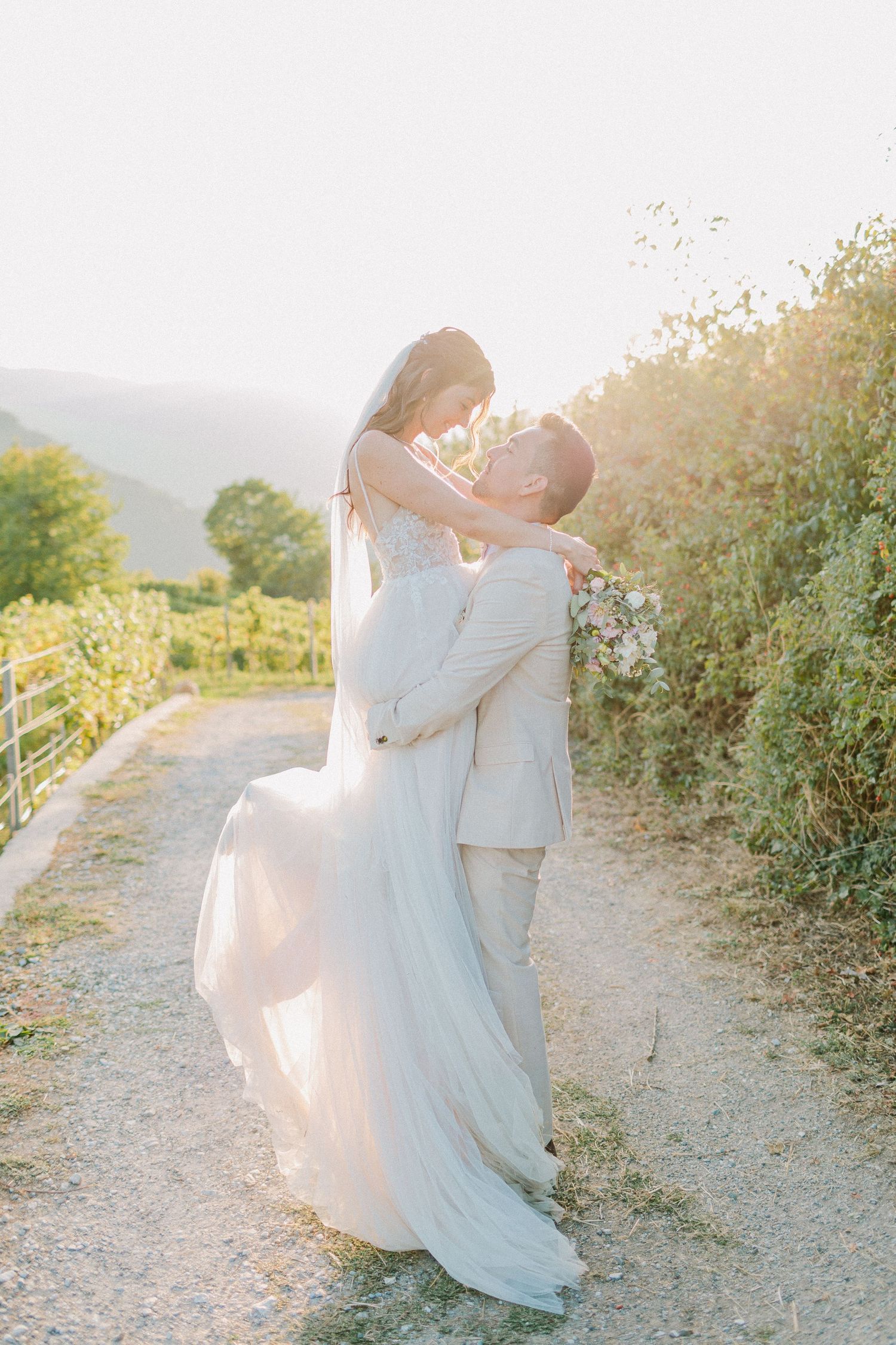 Groom lifting bride in flowing white gown in golden sunset light along a scenic vineyard pathway.