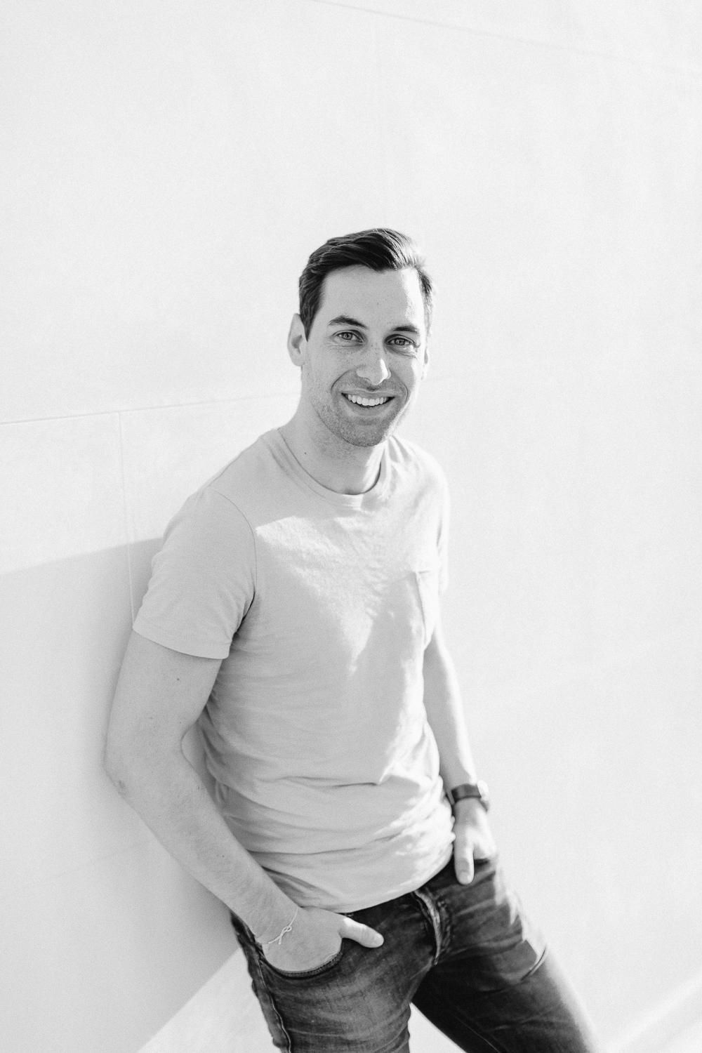 Black and white portrait of a smiling young man in a t-shirt and jeans leaning against a white wall.