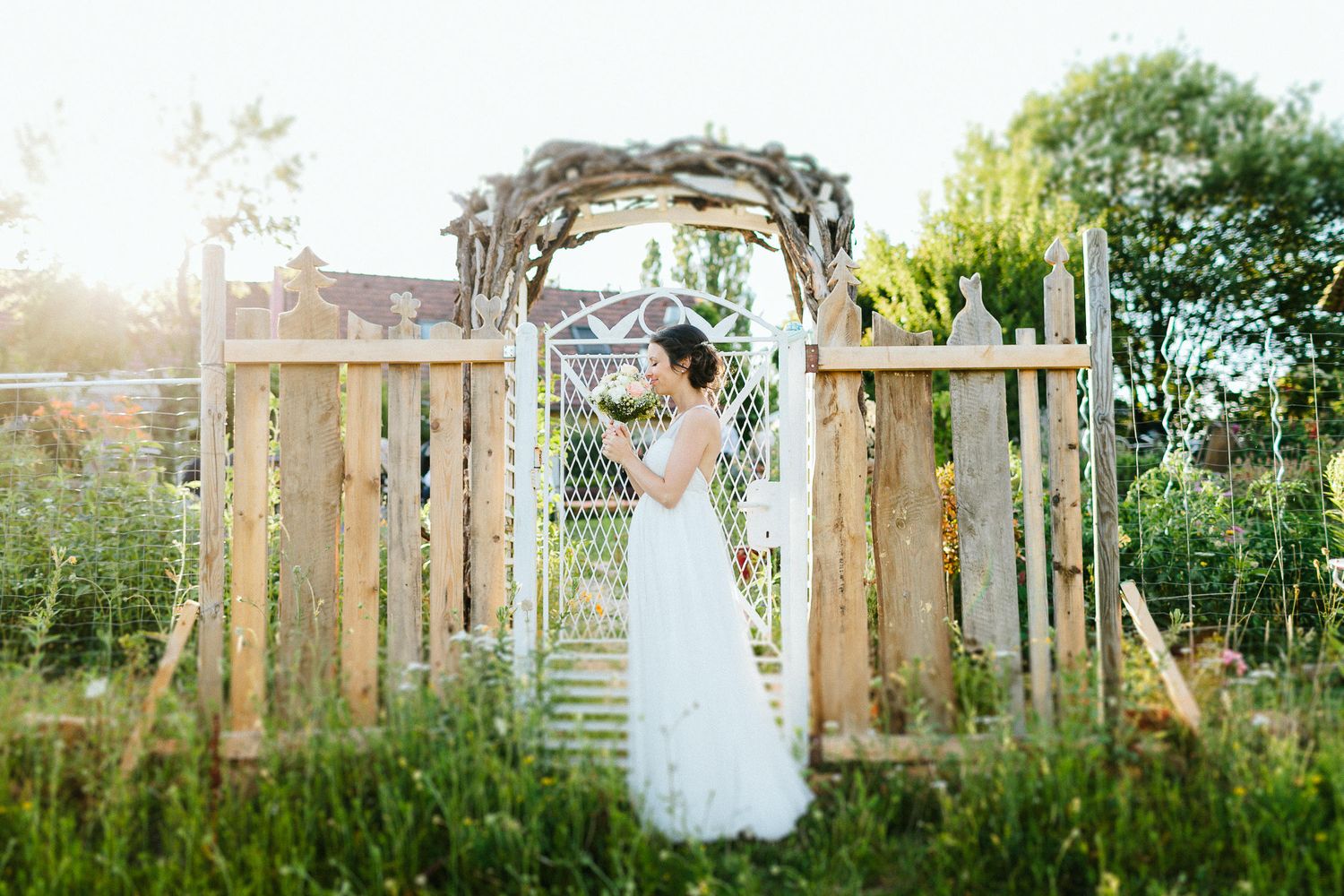 Bride in white lace gown stands beneath a rustic wooden arch gateway surrounded by lush greenery.
