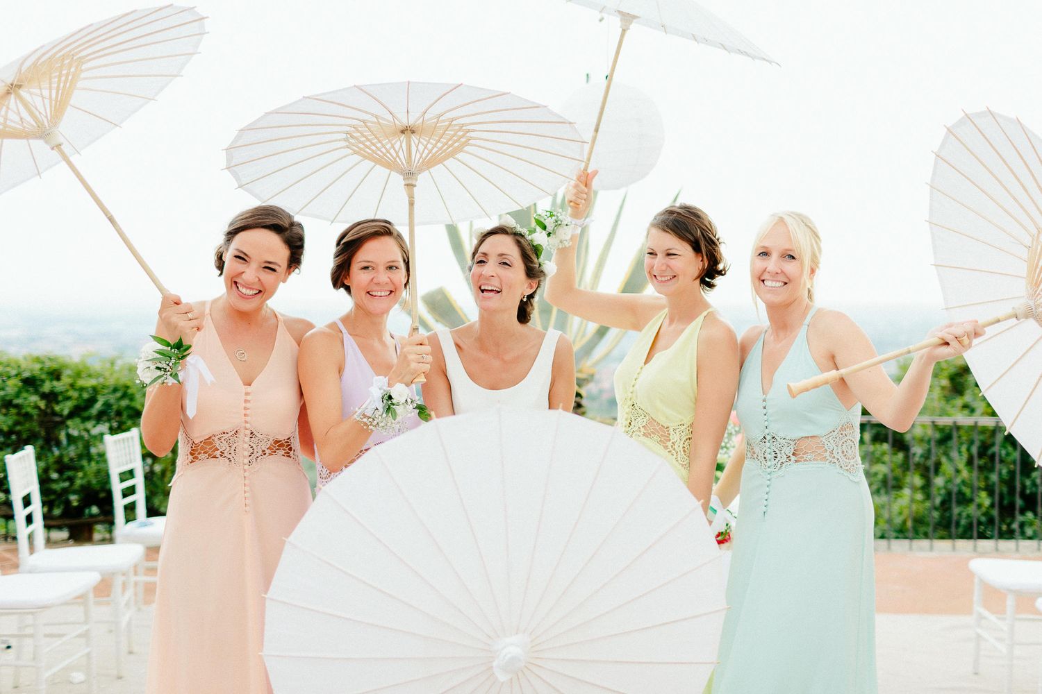 Bride with four bridesmaids in pastel dresses holding white parasols smiling at outdoor wedding celebration.