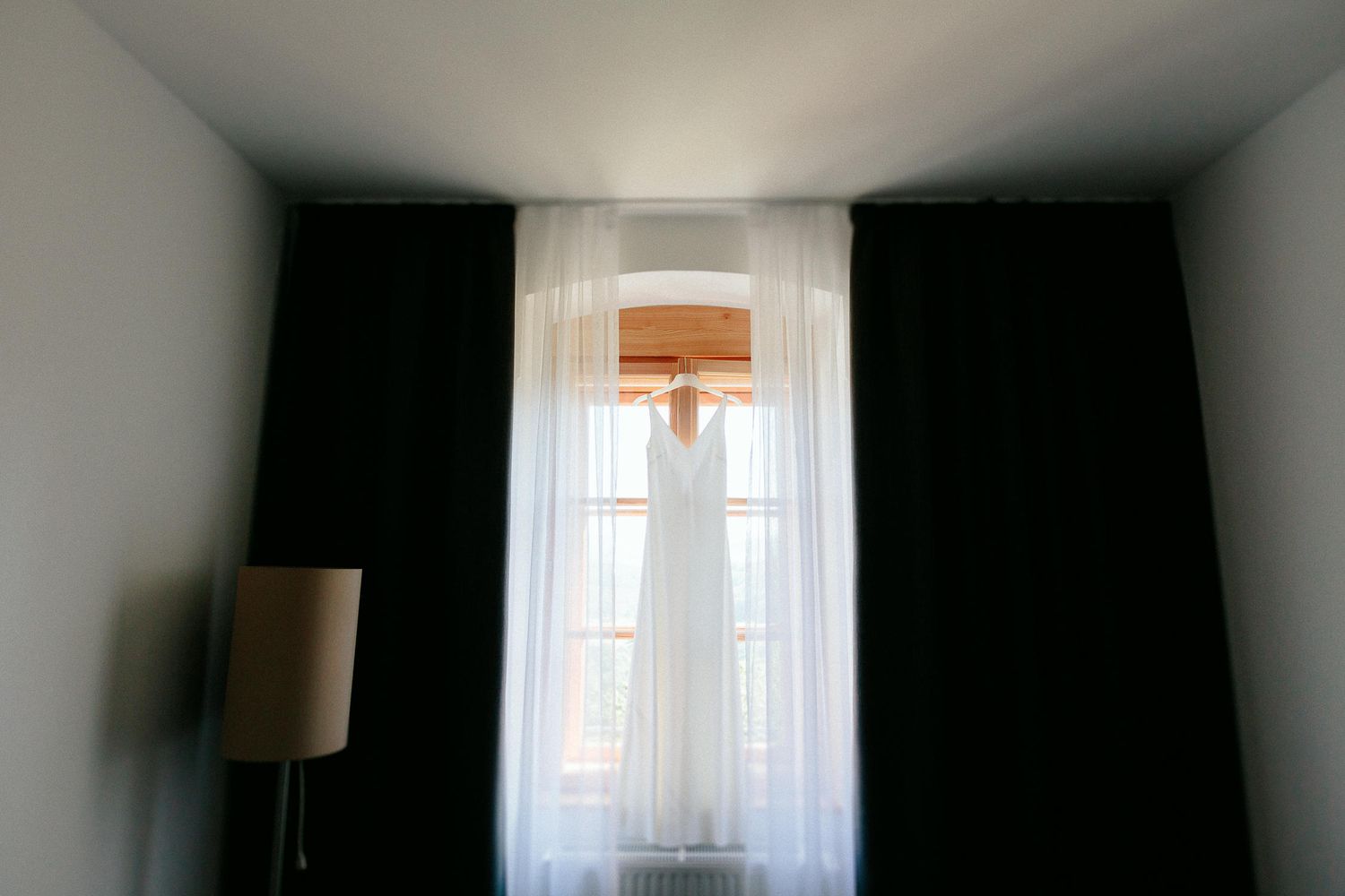 White wedding dress hanging in window between dark curtains with soft natural light filtering through sheers.