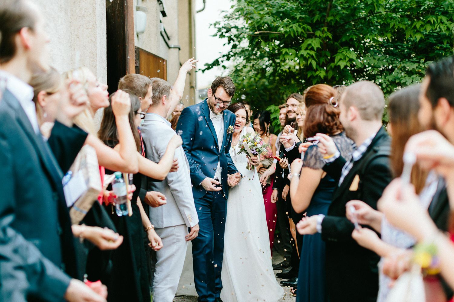 Smiling newlyweds walk through a confetti shower surrounded by cheering wedding guests outside a venue.