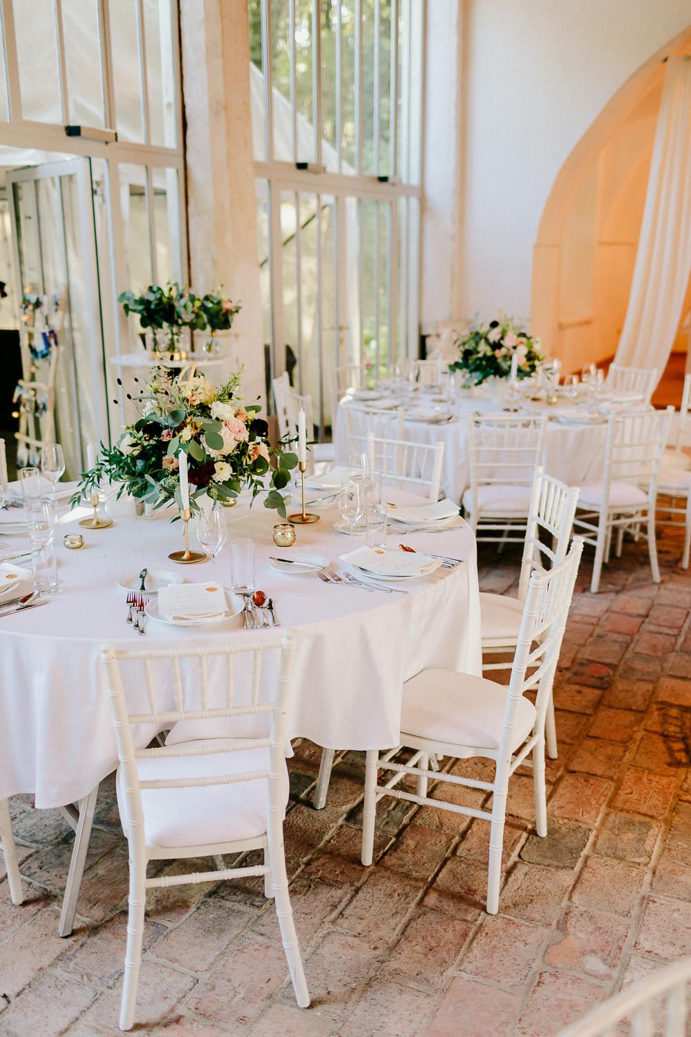 Elegant wedding reception with white chiavari chairs, round tables with floral centerpieces in a bright greenhouse venue.