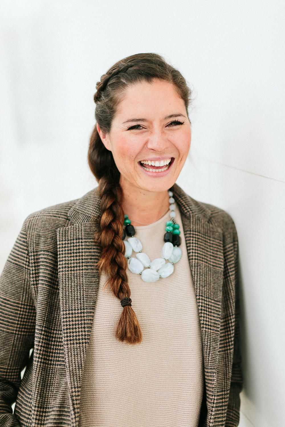 Laughing brunette woman with a long braid wearing a plaid blazer and statement necklace against white wall.