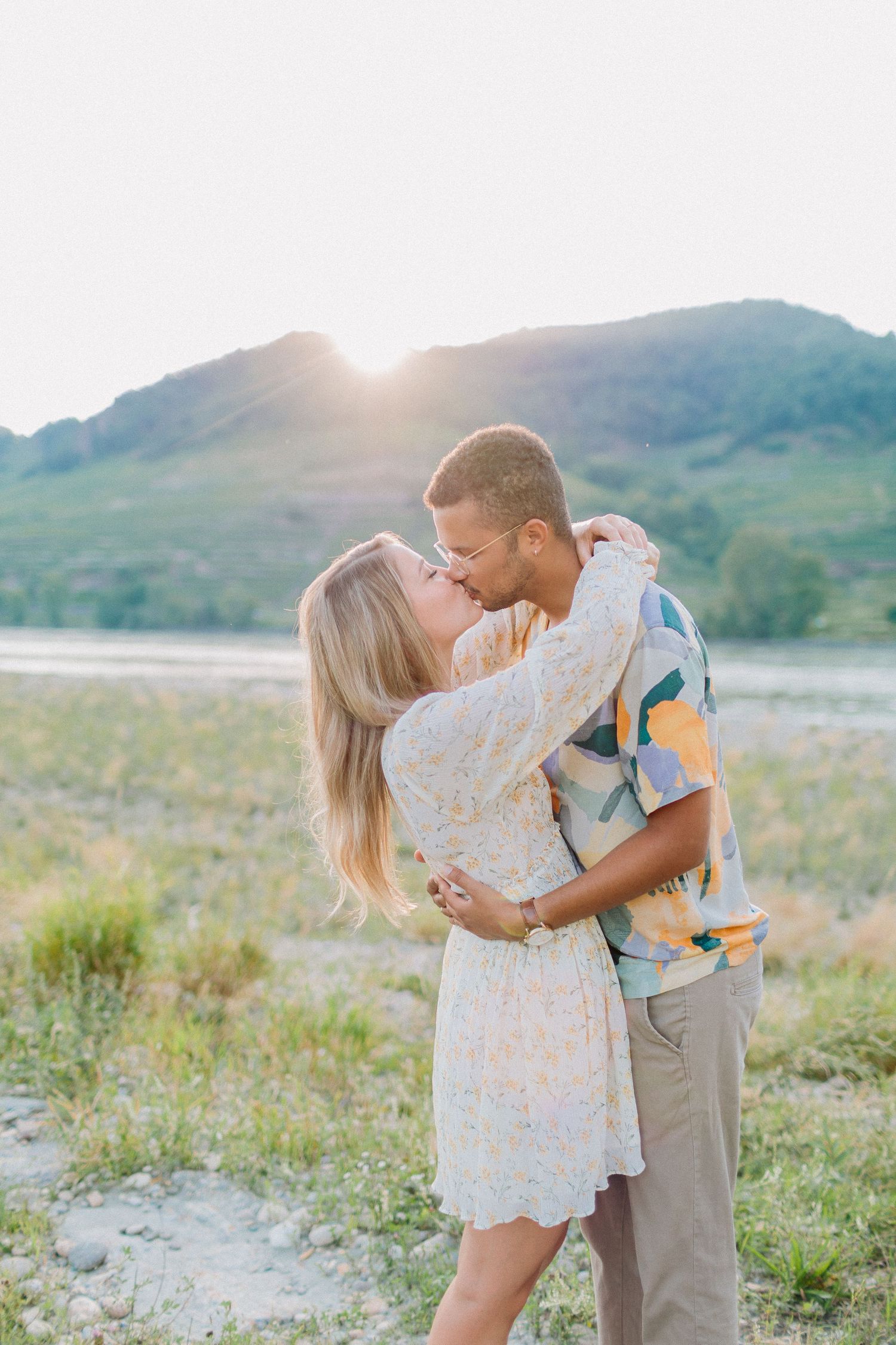 Couple shares a passionate kiss at golden sunset with mountain lake backdrop in casual outdoor engagement session.