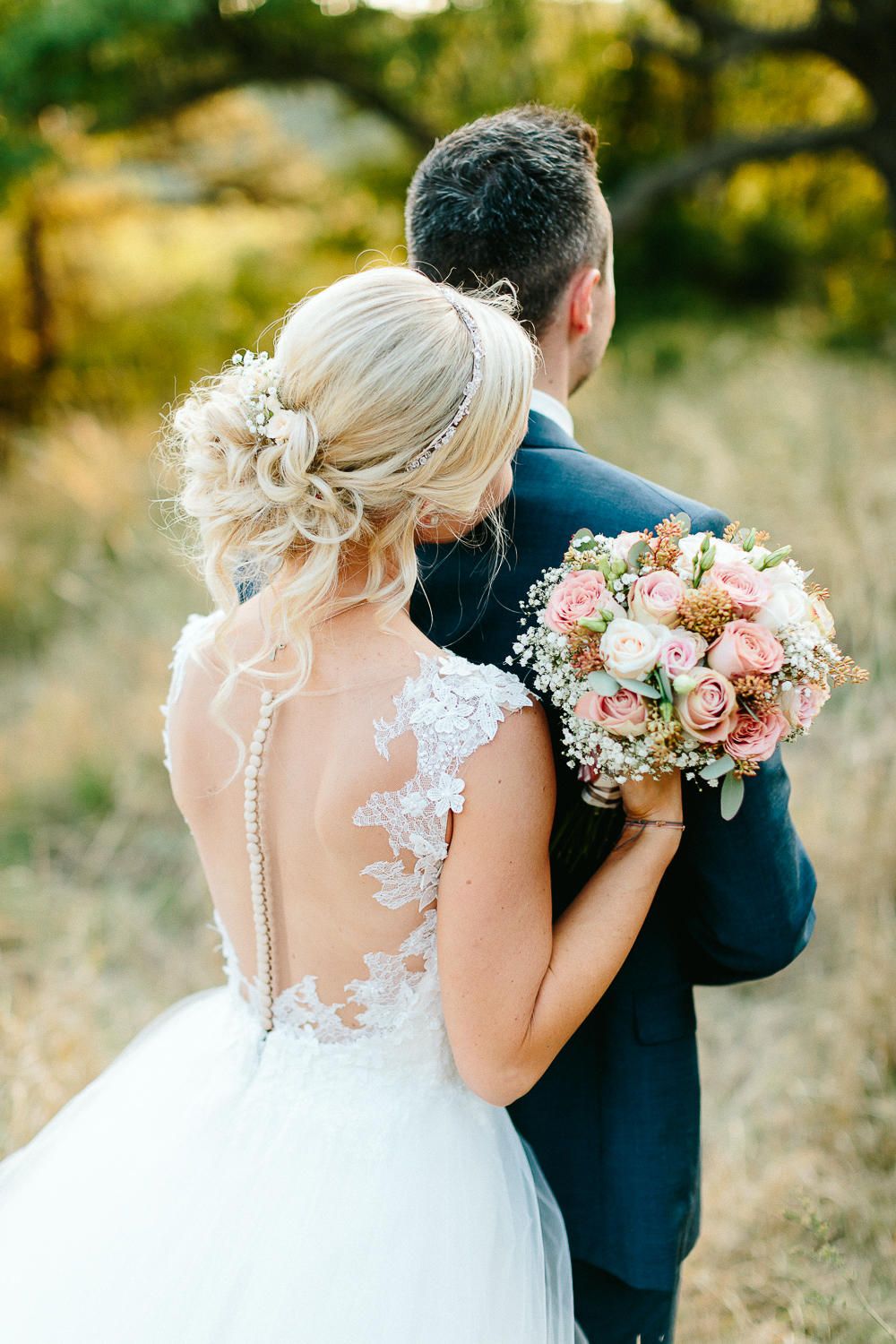 Bride in lace back wedding gown holding pink bouquet walks with groom through golden field.