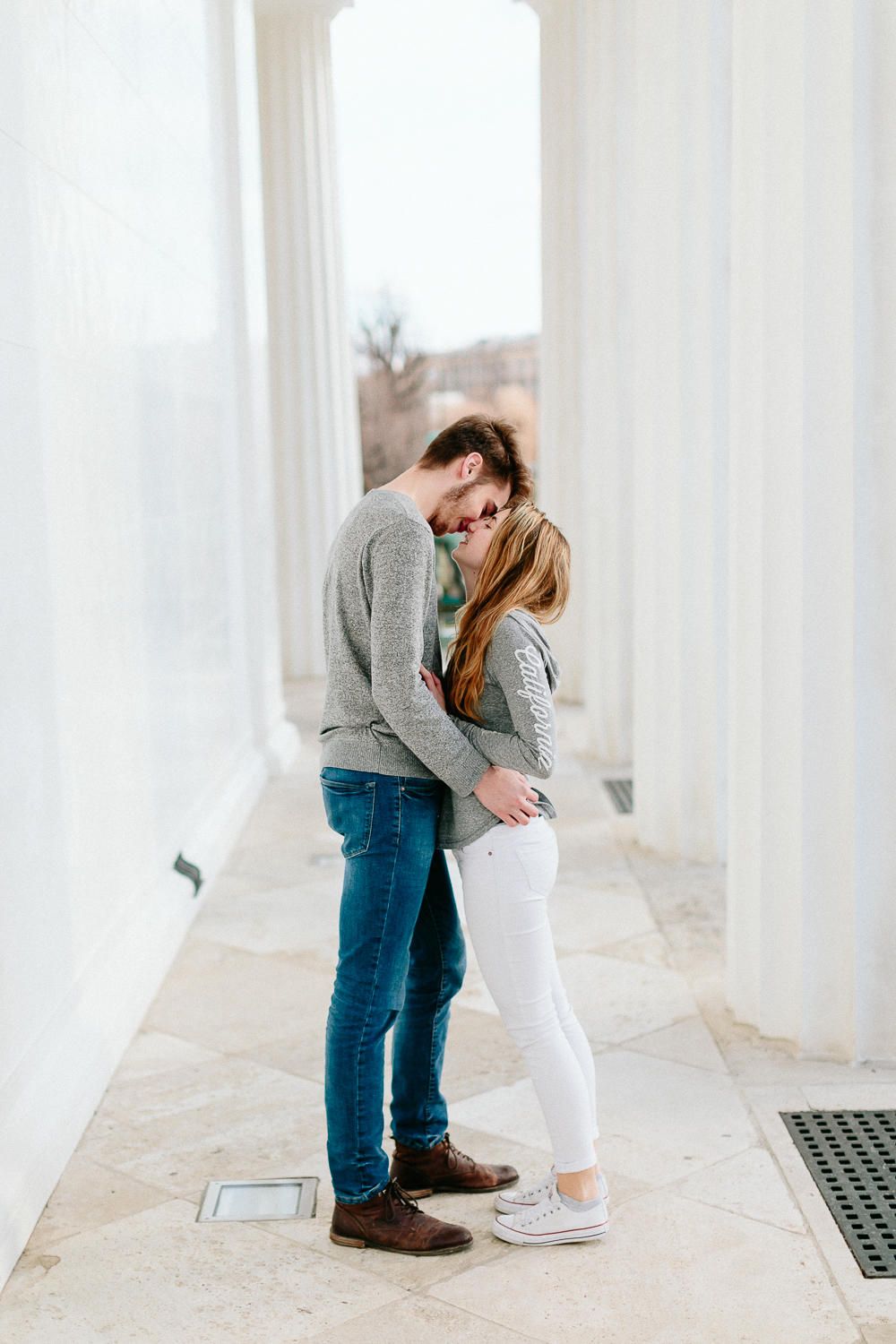 A couple sharing an intimate embrace between white columns in a bright corridor.