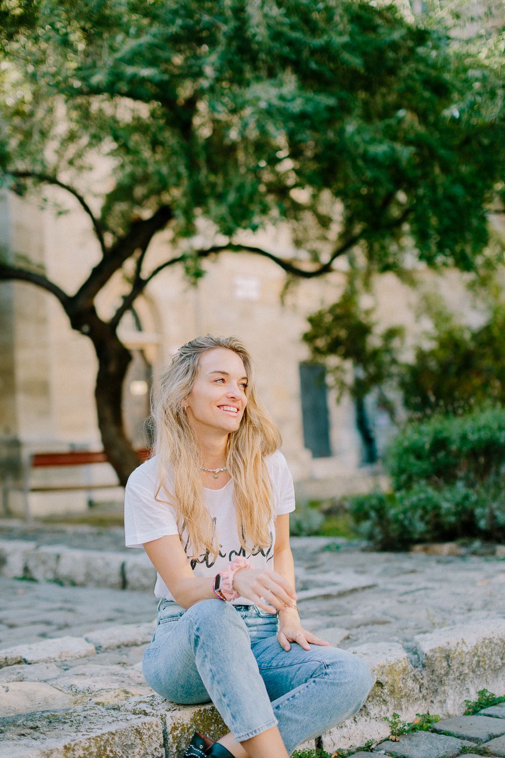 Cheerful blonde woman in white tee and jeans sitting on stone steps near green trees.
