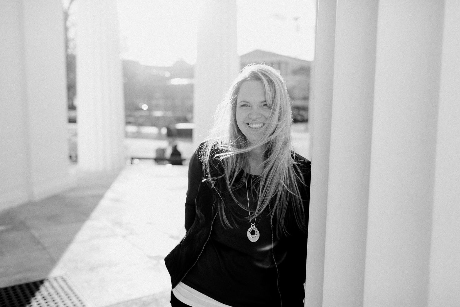 Black and white portrait of smiling blonde woman leaning against white column outdoors.