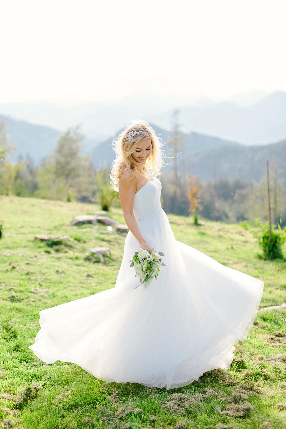 Bride in strapless ball gown twirling in mountain meadow holding white floral bouquet.