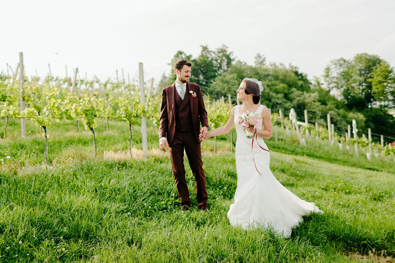 Bride and groom walk hand in hand through lush green vineyard rows on their wedding day.