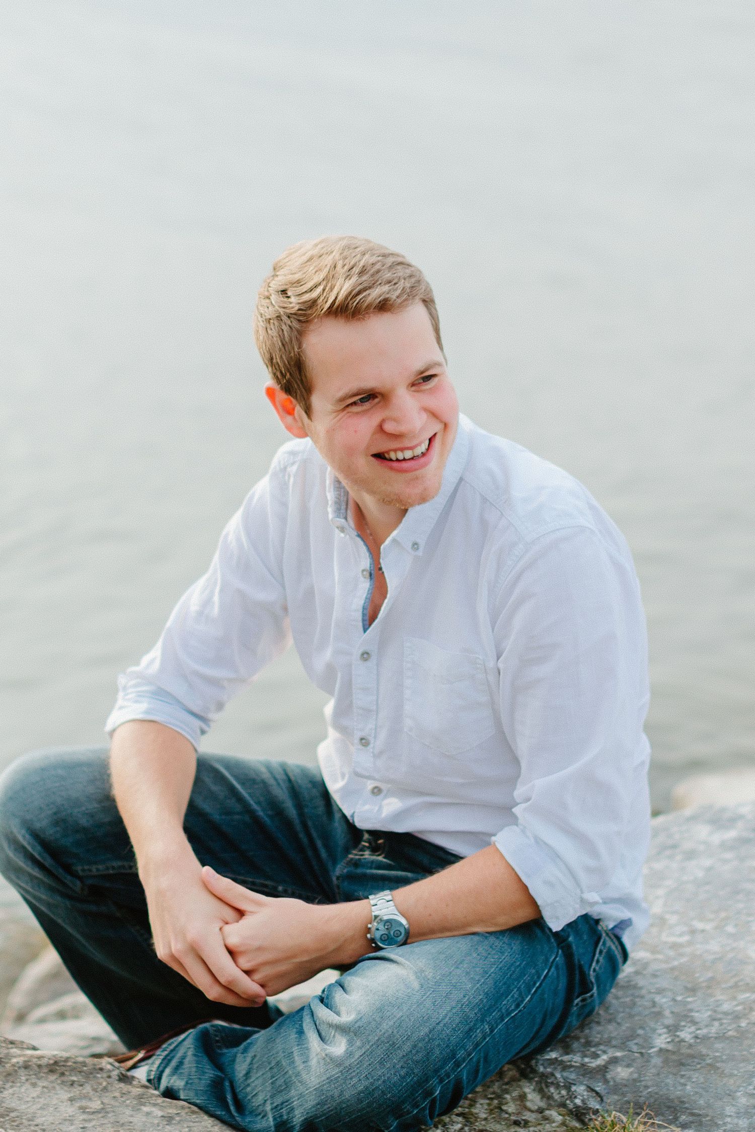 Young man in a white button-up shirt smiling while seated on rocks near calm water.