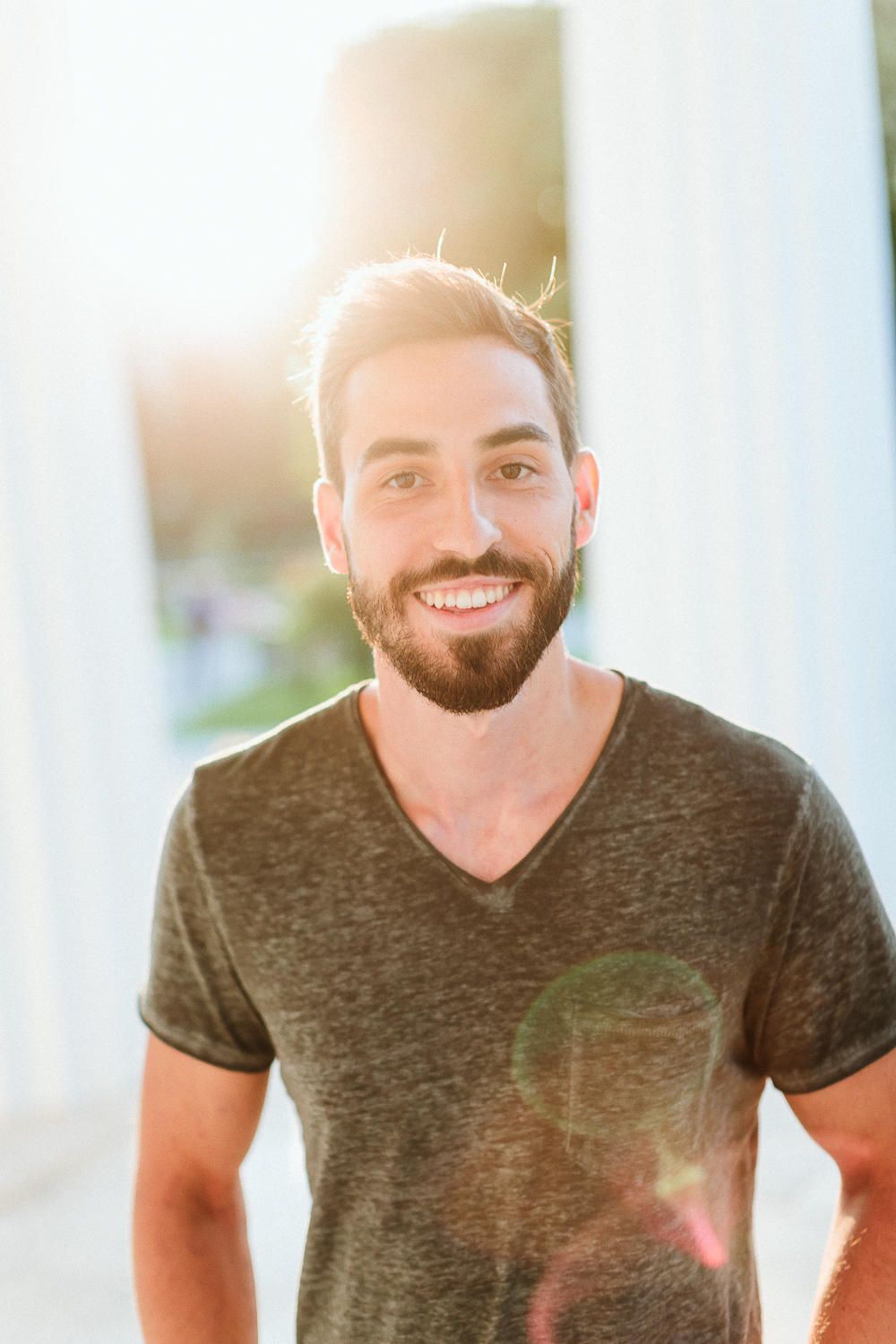 Smiling bearded man in a gray v-neck shirt with bright sunlight glowing behind him.