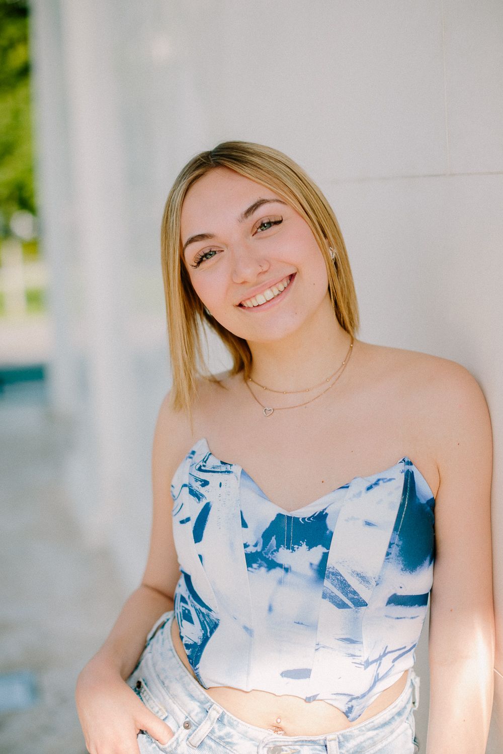 Smiling young blonde woman in a blue and white strapless crop top leaning against a white wall outdoors.