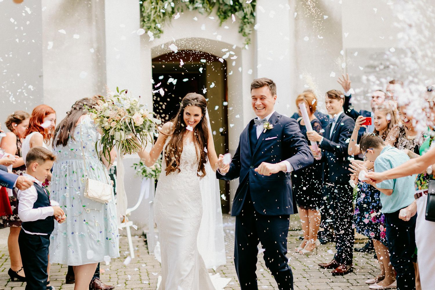 Newlyweds exit a church ceremony as guests shower them with white confetti in celebration.
