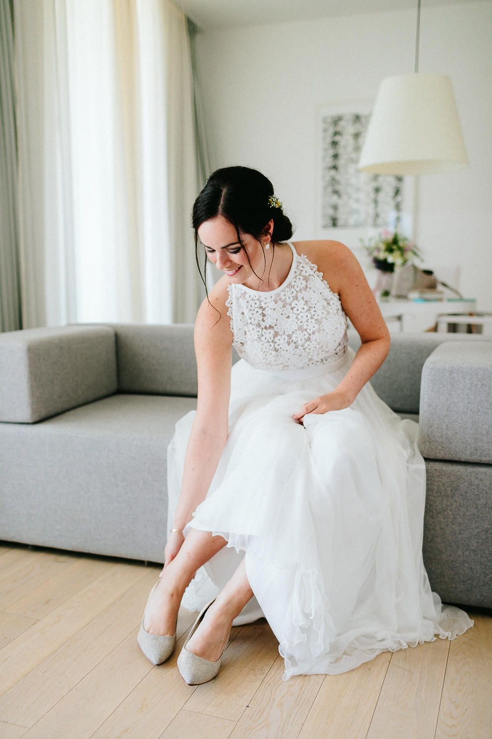 Elegant bride in lace halter wedding dress sitting on grey sofa adjusting her white heels indoors.