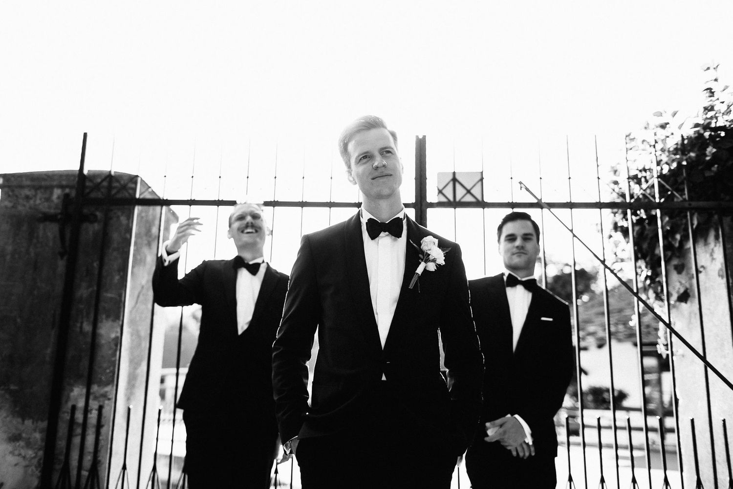 Three groomsmen in black tuxedos pose confidently in front of iron gates in black and white photo.