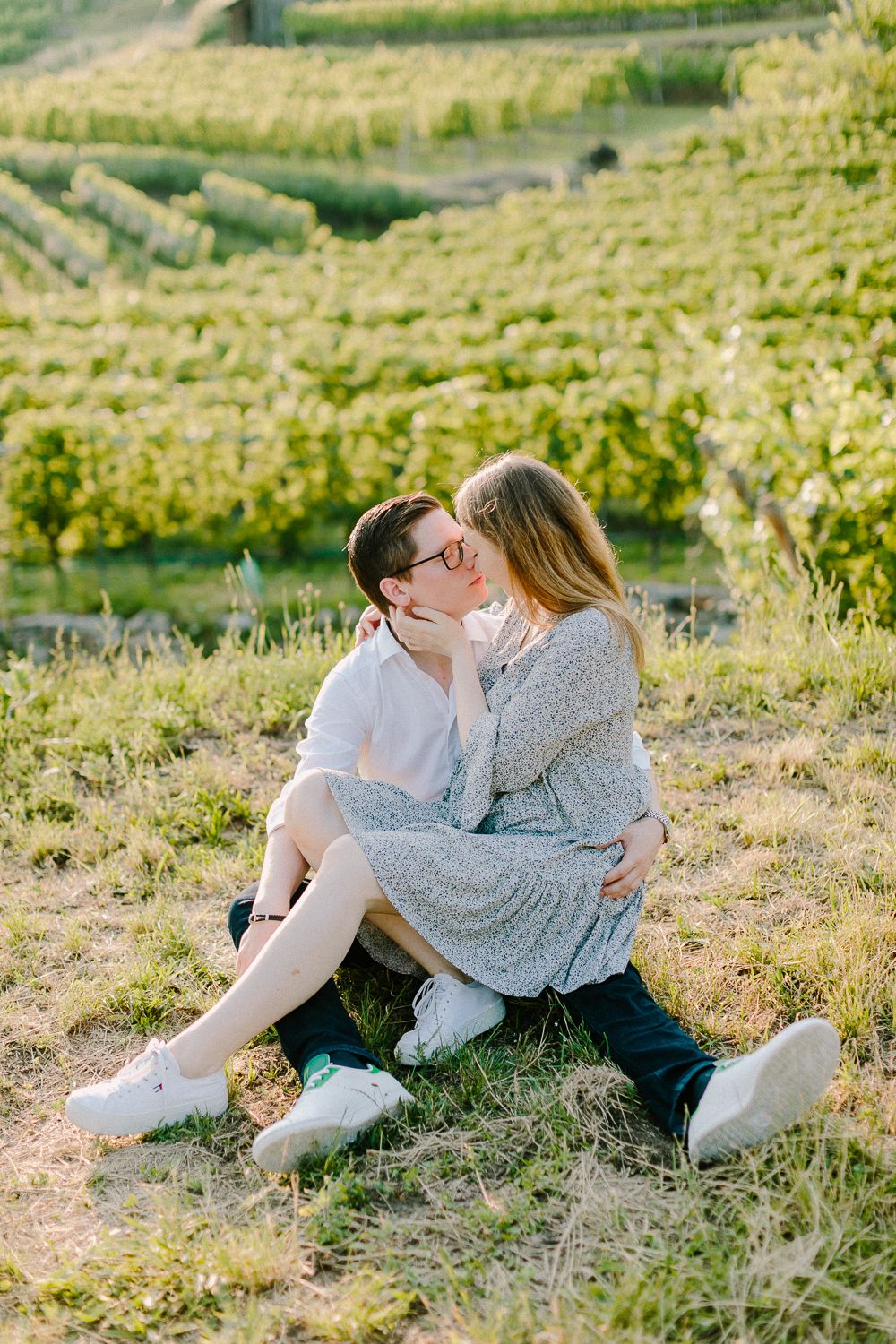Couple sharing a tender kiss while sitting on a grassy hillside overlooking lush green vineyards.