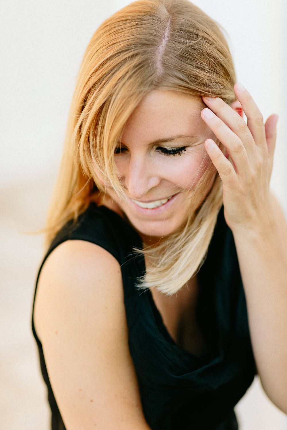 Smiling blonde woman in a black top touching her hair in soft natural light.