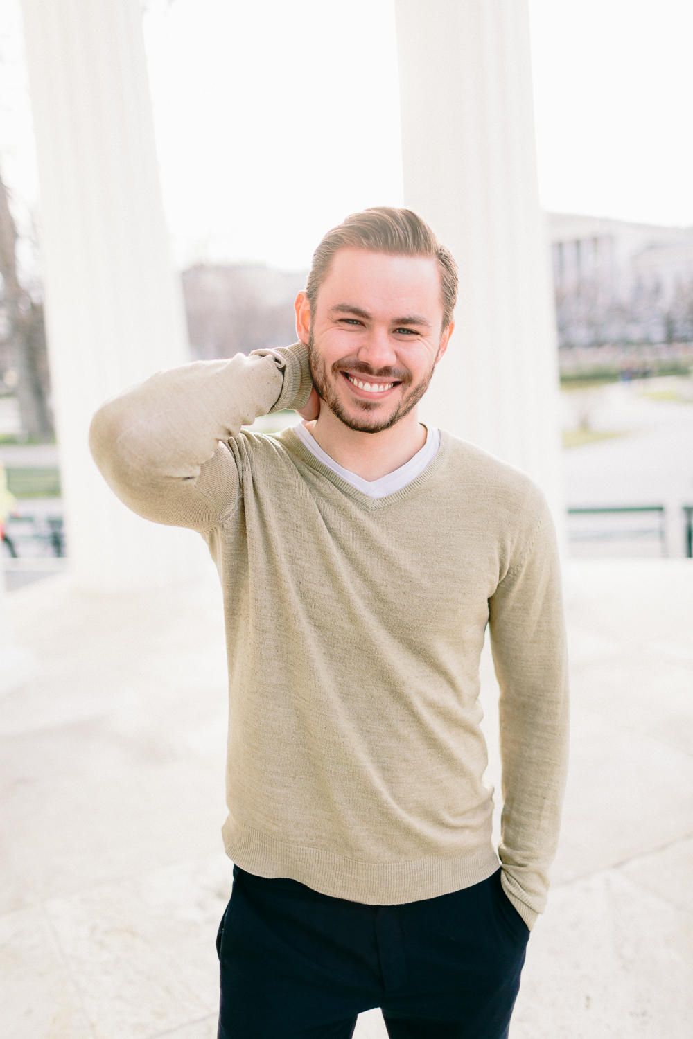 Cheerful bearded young man in a beige sweater laughing while touching the back of his neck outdoors.