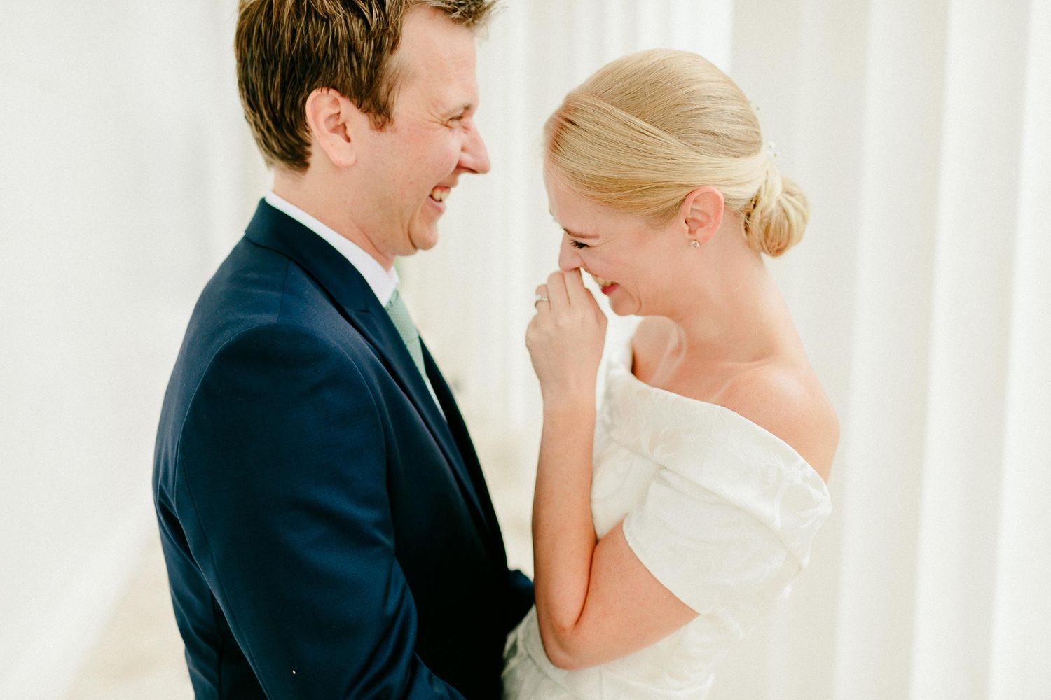 Laughing bride in off-shoulder gown shares a joyful moment with smiling groom against white backdrop.