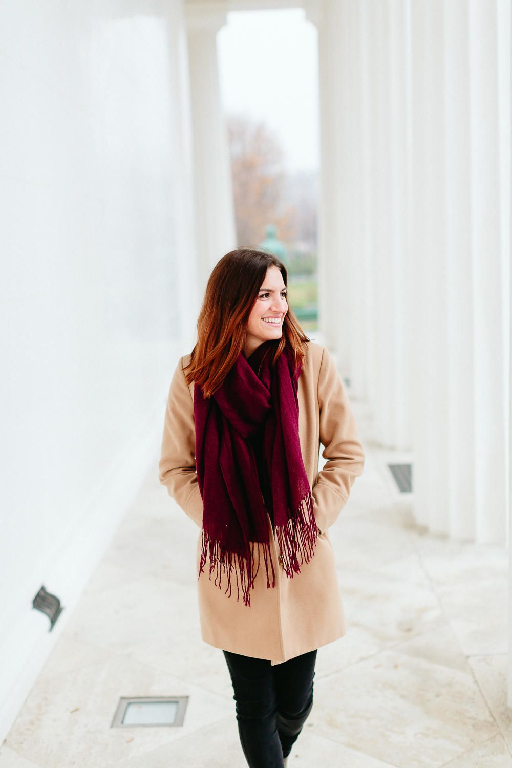 Smiling woman in beige coat and burgundy fringe scarf walking between white columns outdoors.