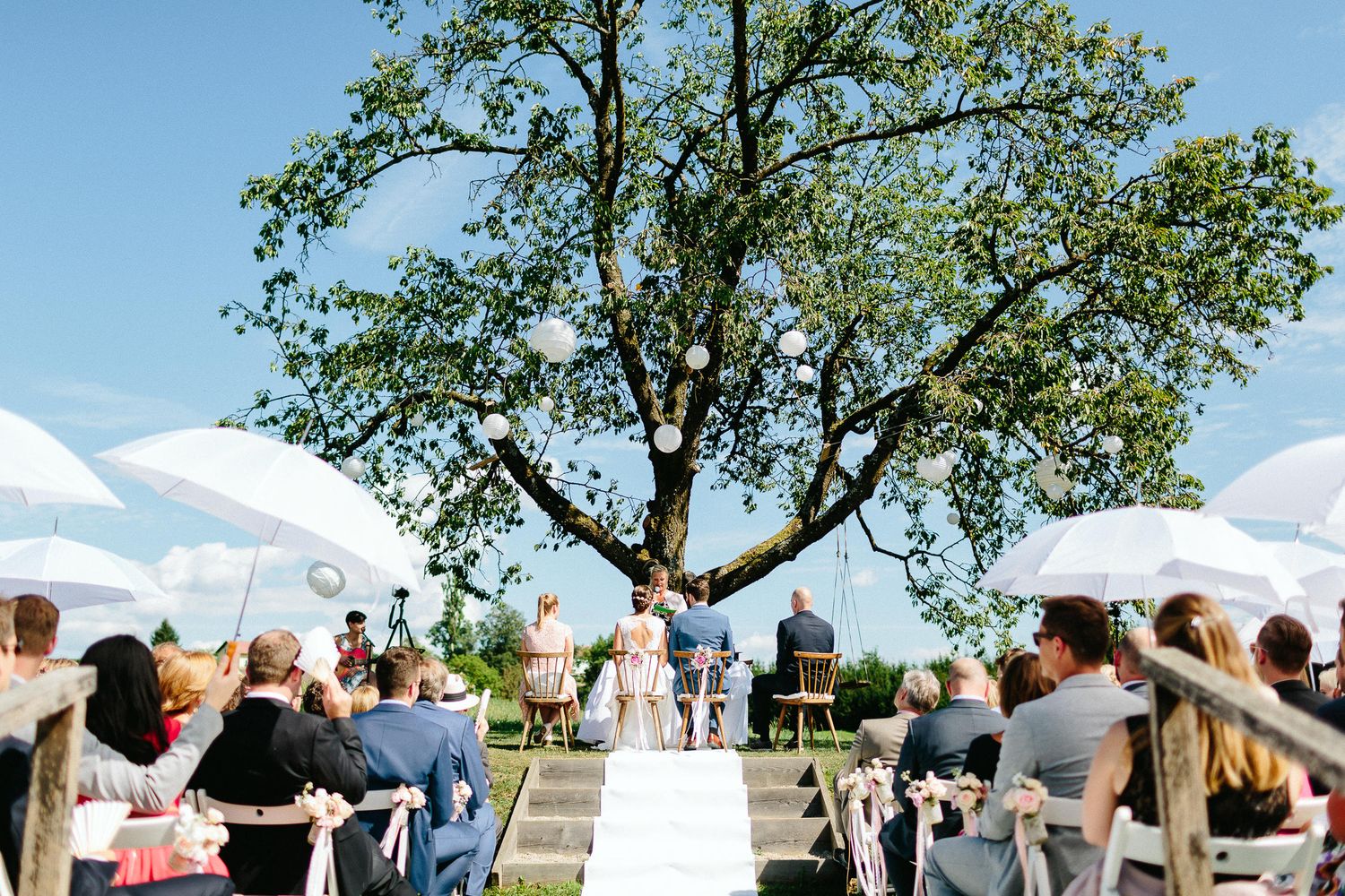 Outdoor wedding ceremony with guests seated facing altar under large tree with white paper lanterns on sunny day.