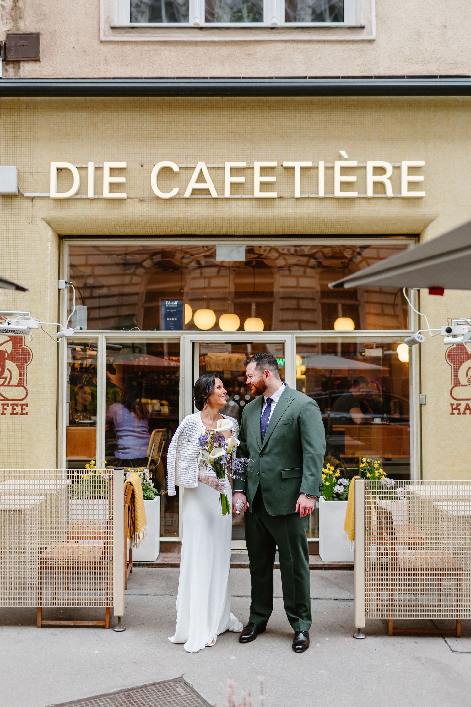 Newlyweds posing together outside Die Cafetiere cafe, bride in white gown, groom in green suit.