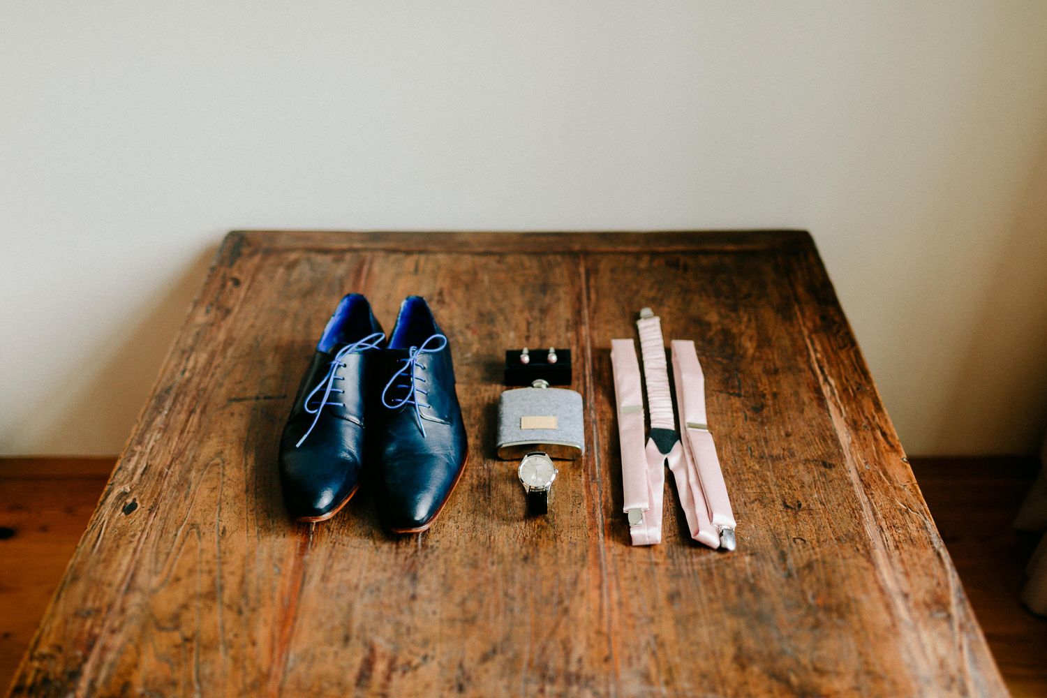 Groom wedding accessories flat lay with navy dress shoes, tie, cologne and cufflinks on wood table.