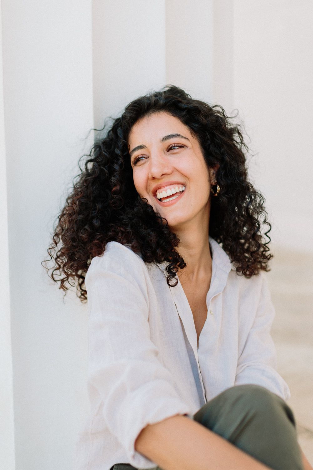 Happy young woman with curly dark hair laughing, wearing white linen shirt outdoors.
