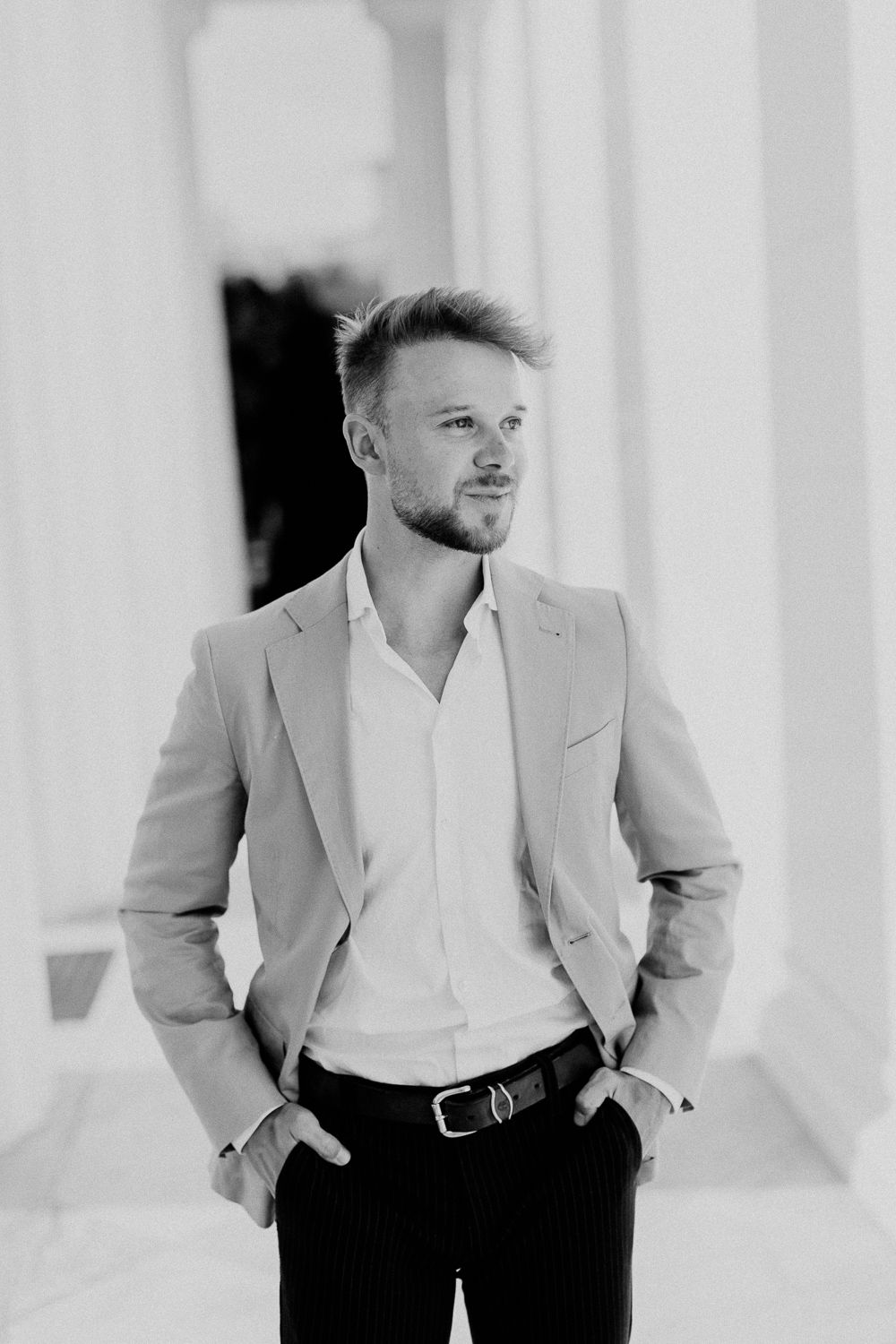 Stylish man in blazer and white shirt standing between columns in black and white portrait.
