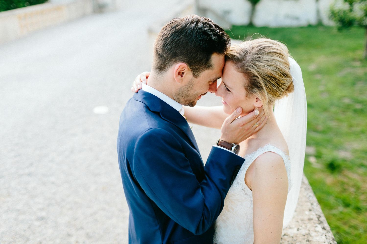 Groom in navy suit tenderly holds bride's face as they share an intimate moment outdoors.