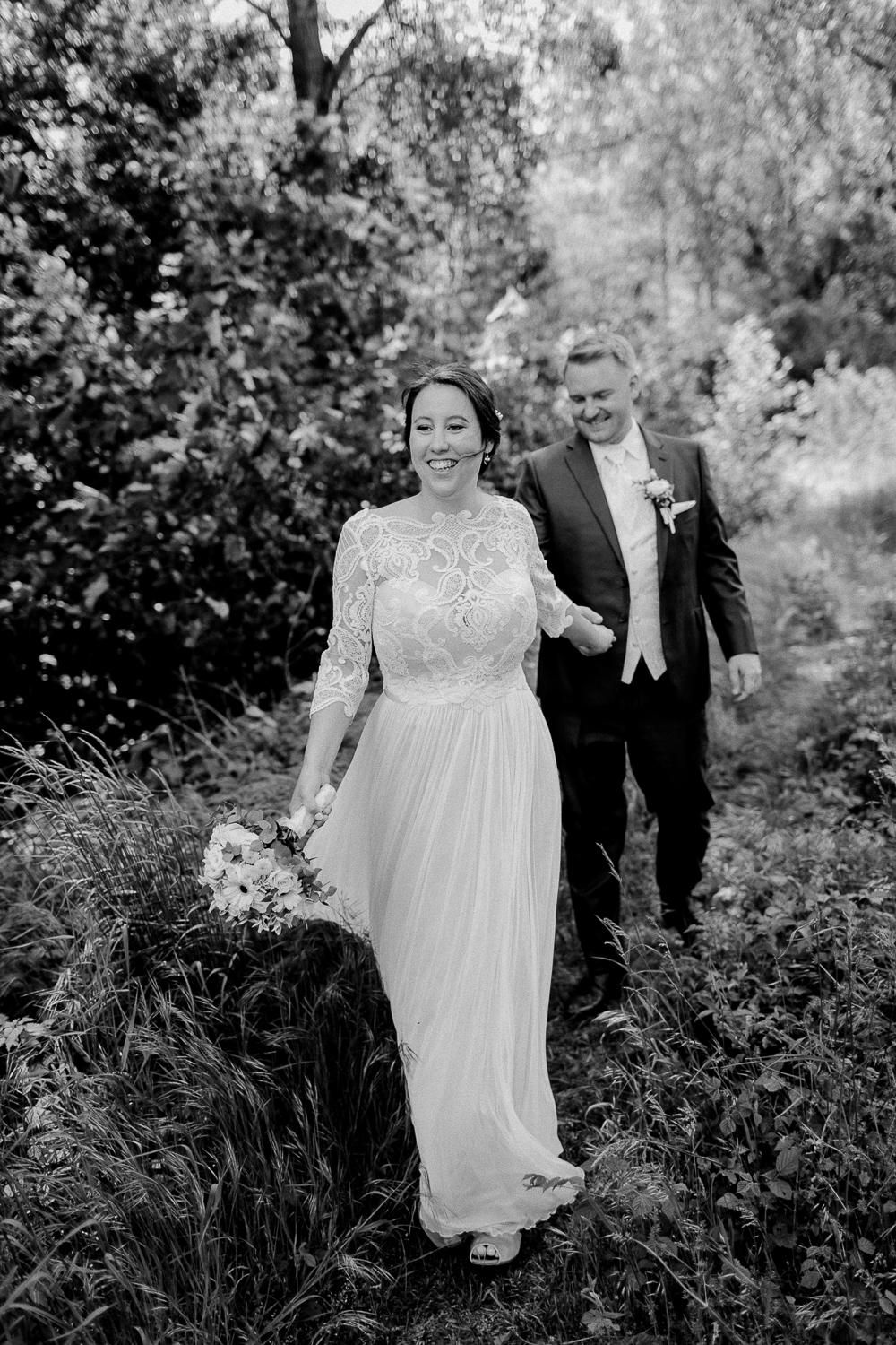 Black and white photo of smiling bride in lace gown walking with groom through lush garden greenery.