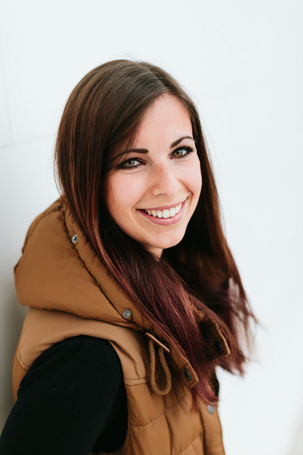 Young woman with long brown hair smiling brightly, wearing a tan vest outdoors.