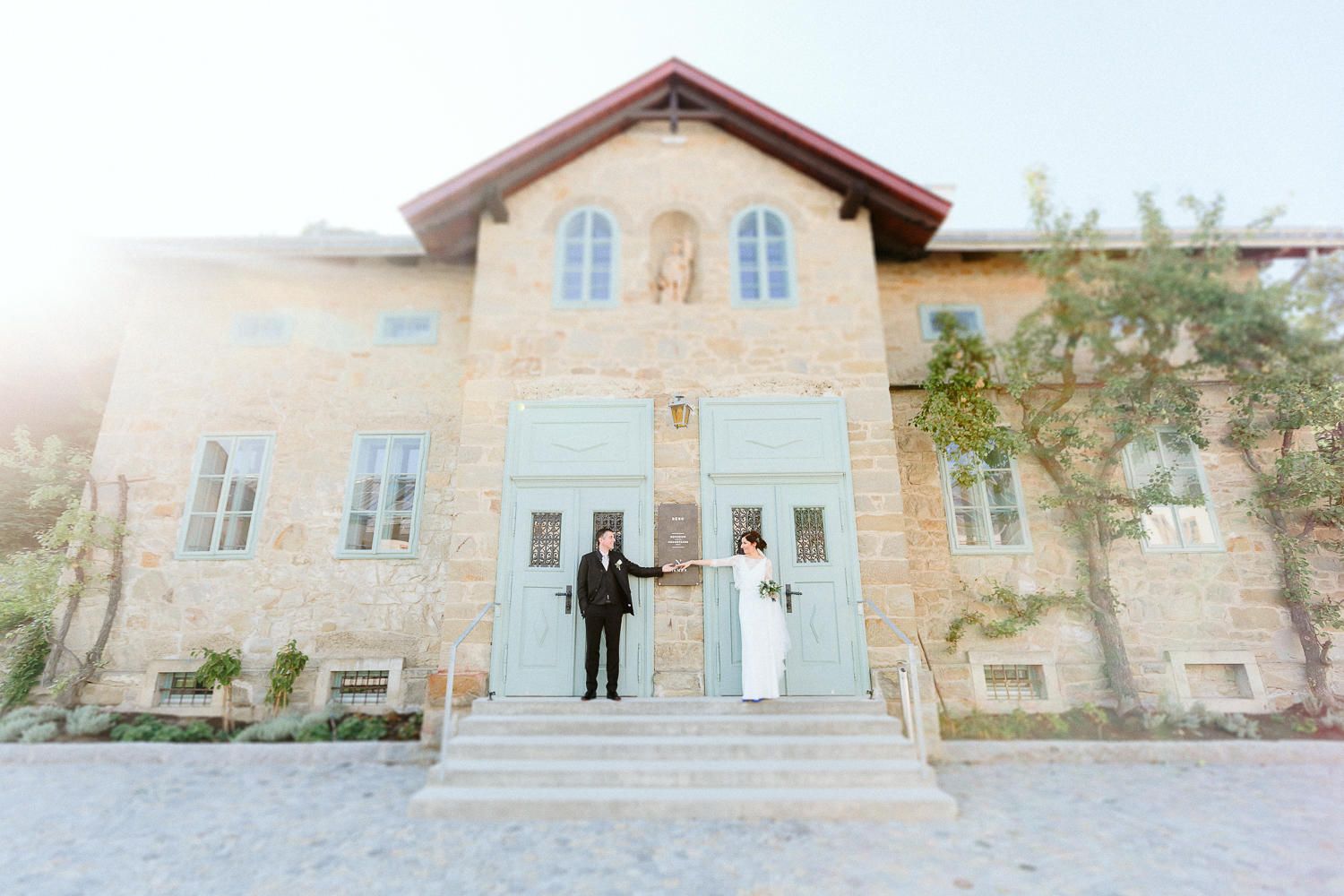 Bride and groom holding hands on steps of rustic stone building with light blue doors on wedding day.