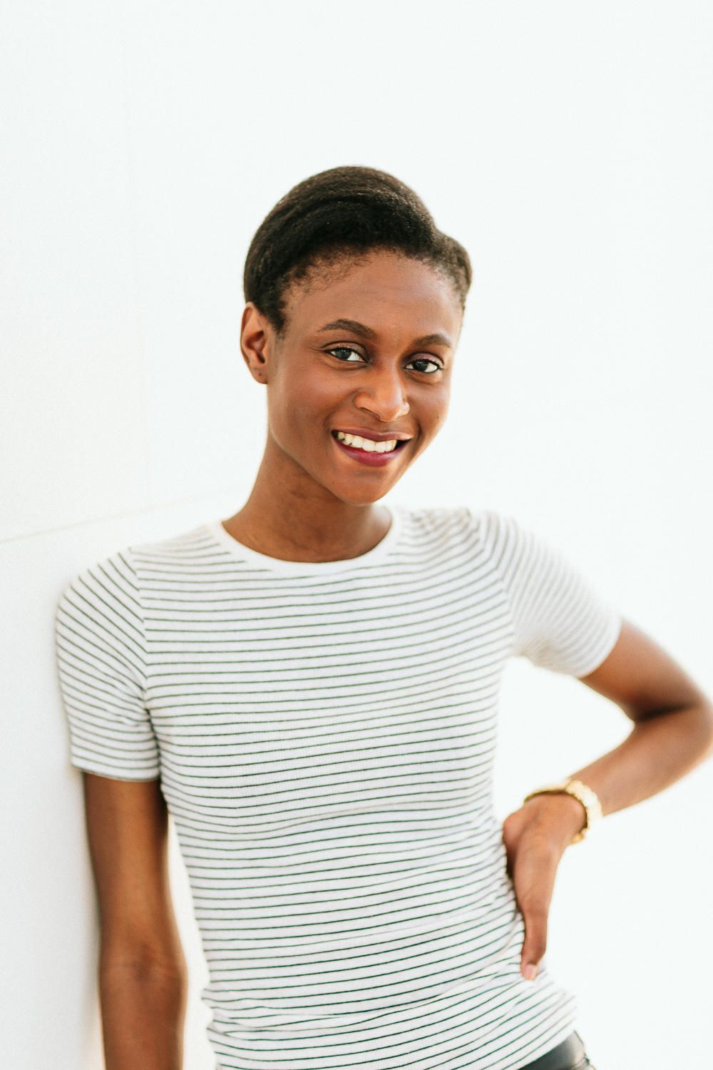 Smiling Black woman in striped white t-shirt leaning against a white wall wearing gold bracelet.