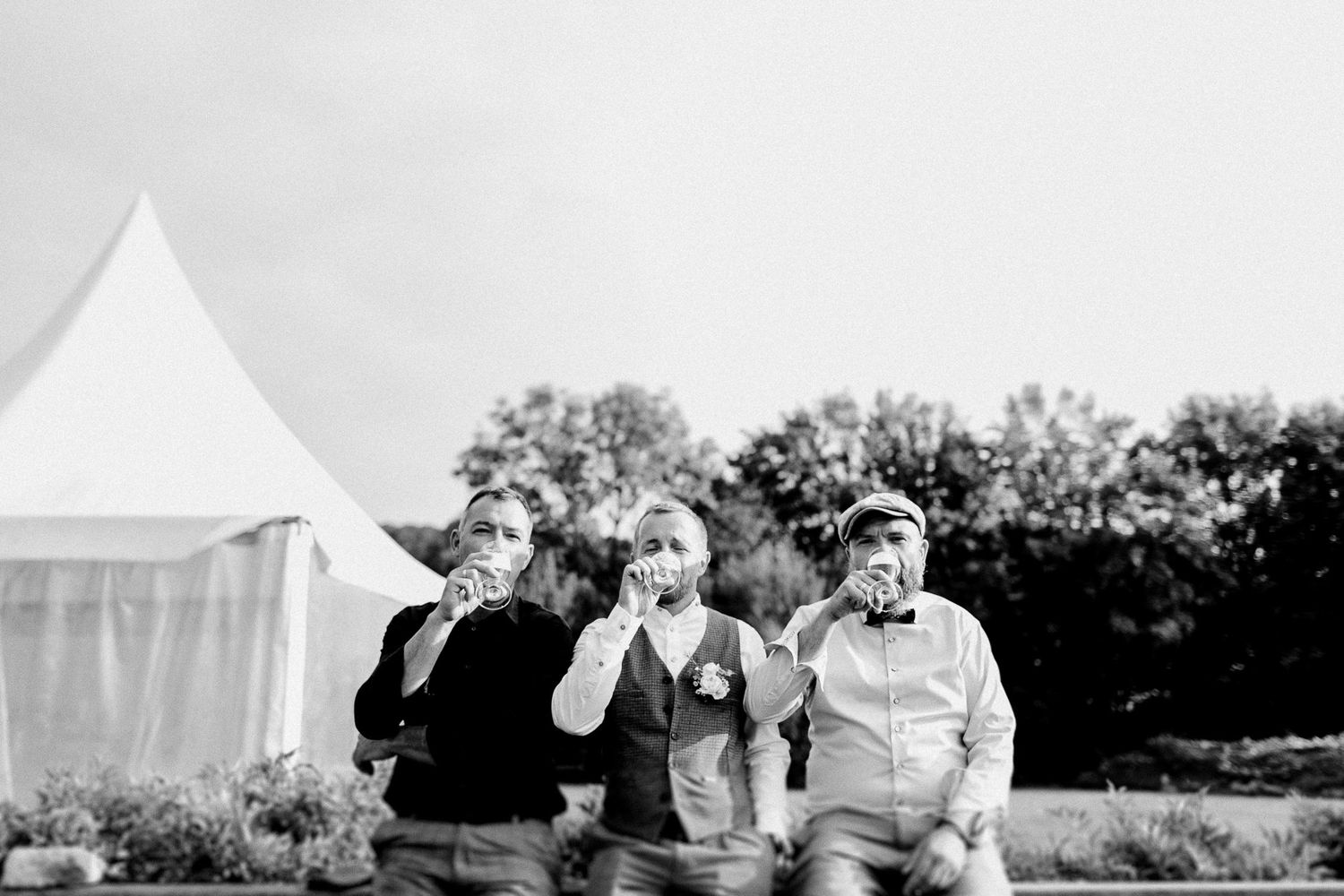 Three wedding guests sit outdoors drinking in black and white photo near a white tent.