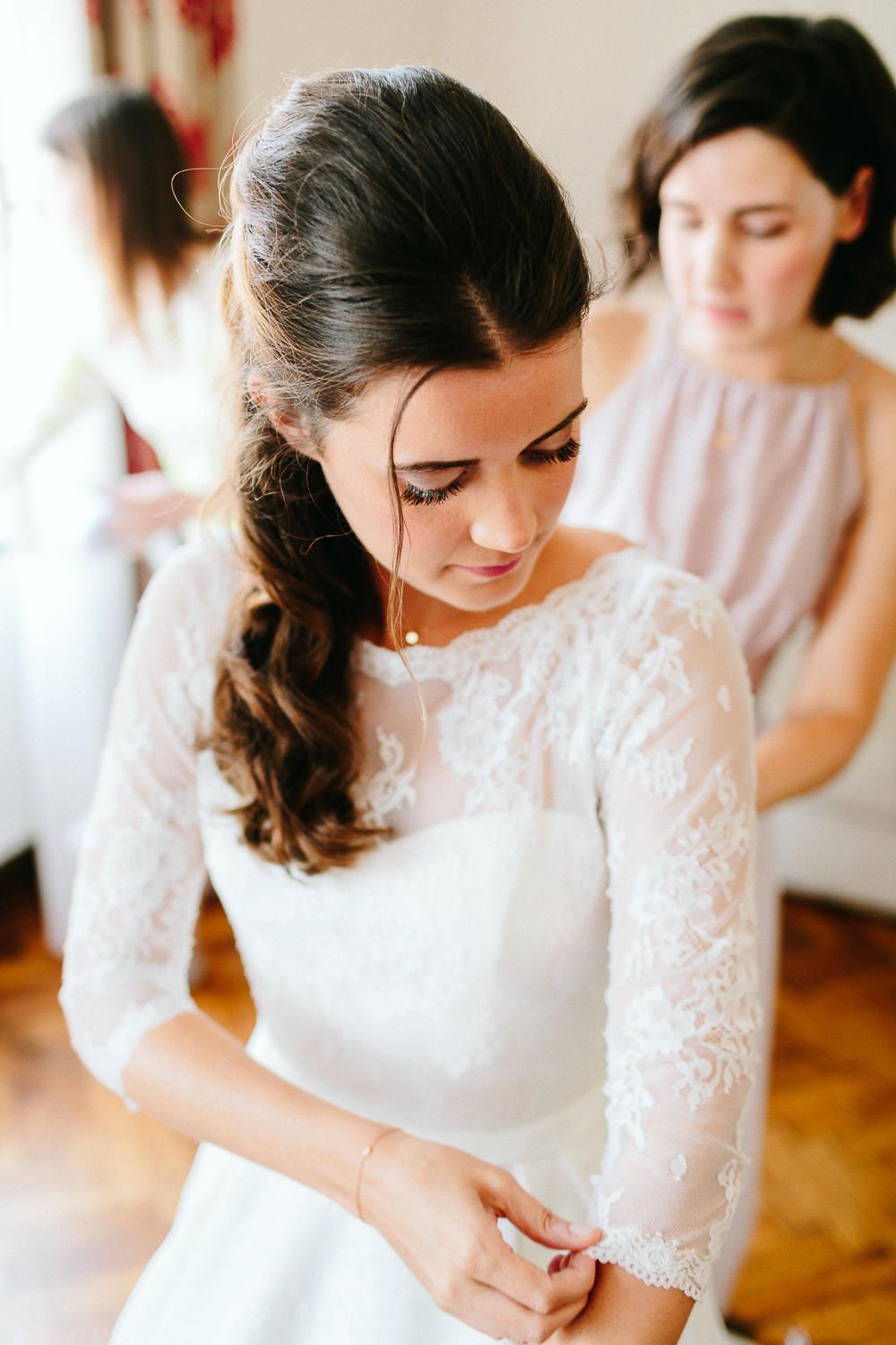 Bride in lace wedding gown adjusting her sleeve while bridesmaid assists with dress in bright room.